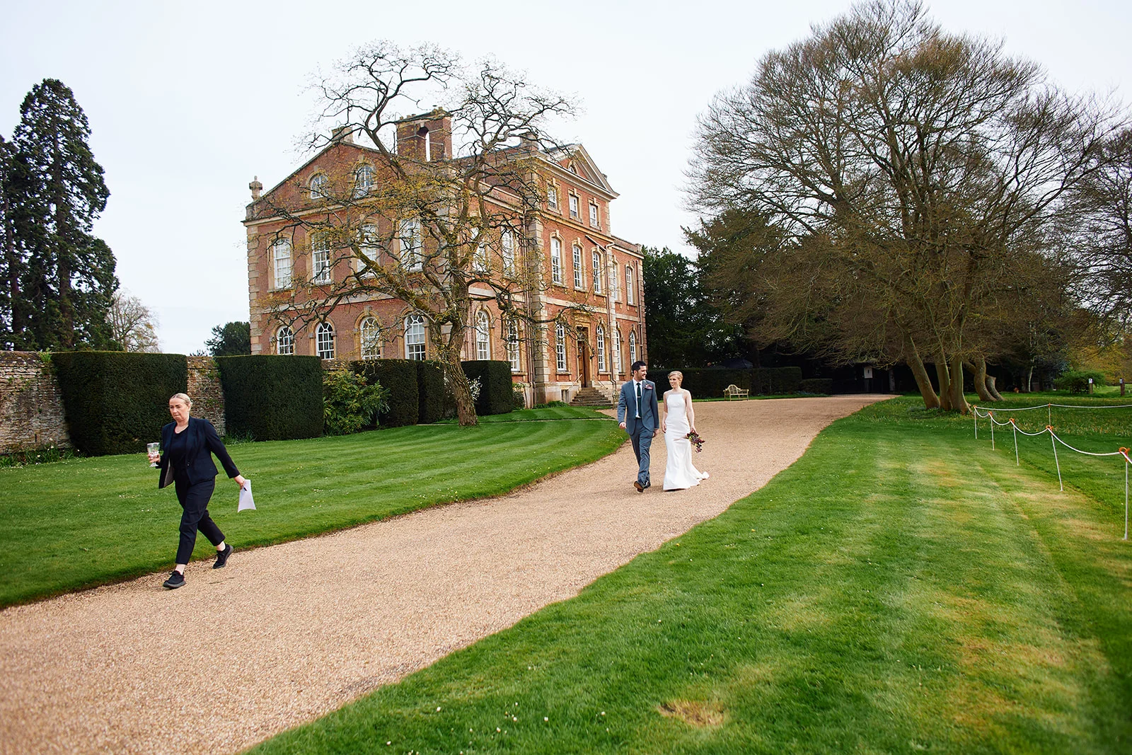 A bride and groom walk hand in hand along a gravel path in front of a grand manor house while a staff member walks on the lawn nearby.