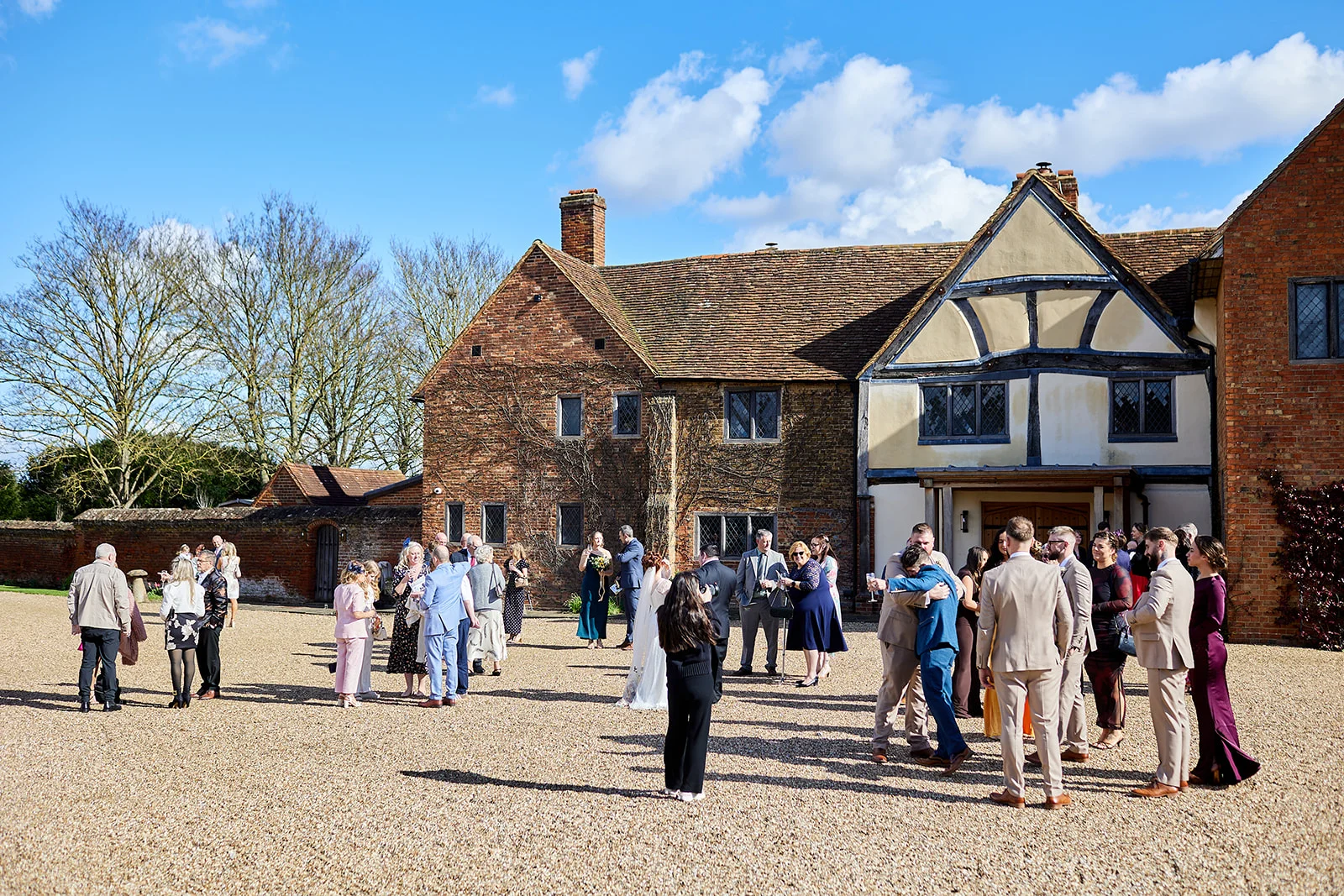 Wedding guests mingle and enjoy drinks in a gravel courtyard outside a historic brick and timber-framed manor house on a sunny day.