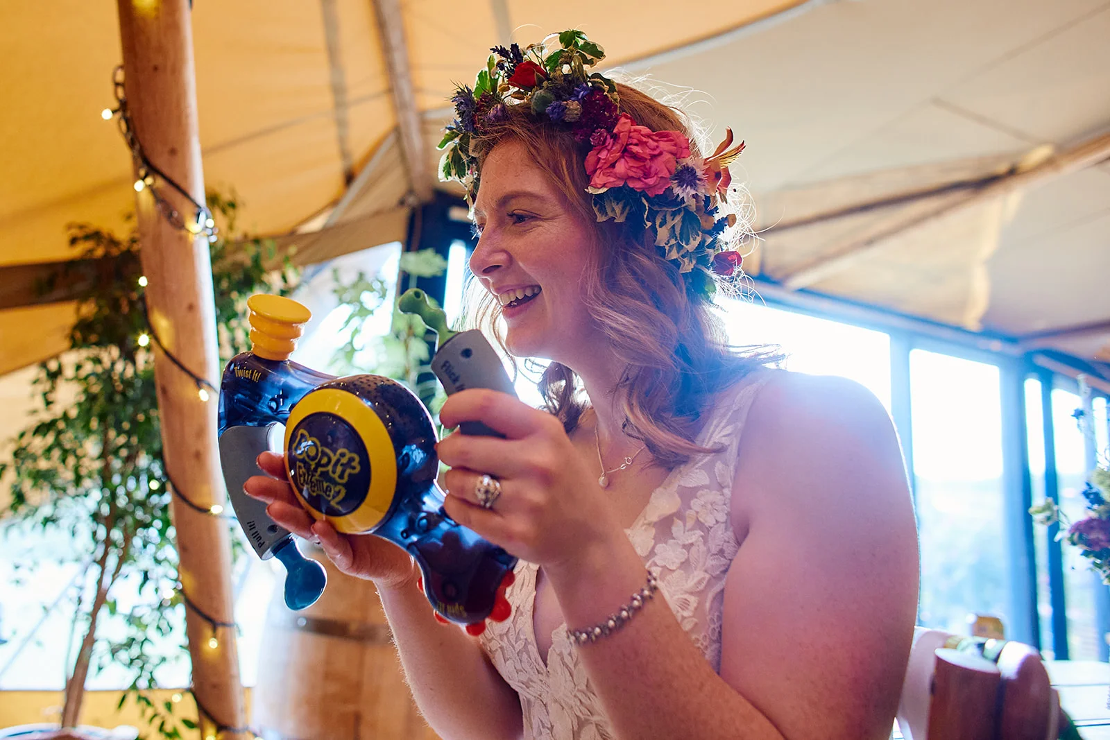 A laughing bride wearing a fresh floral crown and a lace V-neck dress plays with a toy inside a marquee wedding reception.
