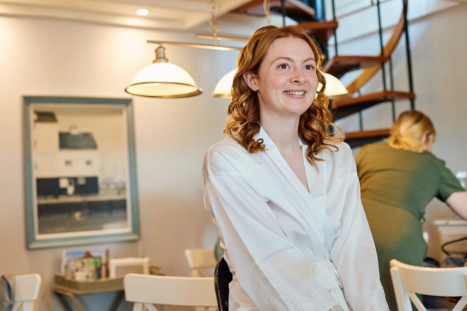 A smiling red-haired bride wearing a white getting ready robe looks off to the side indoors during wedding morning preparations.