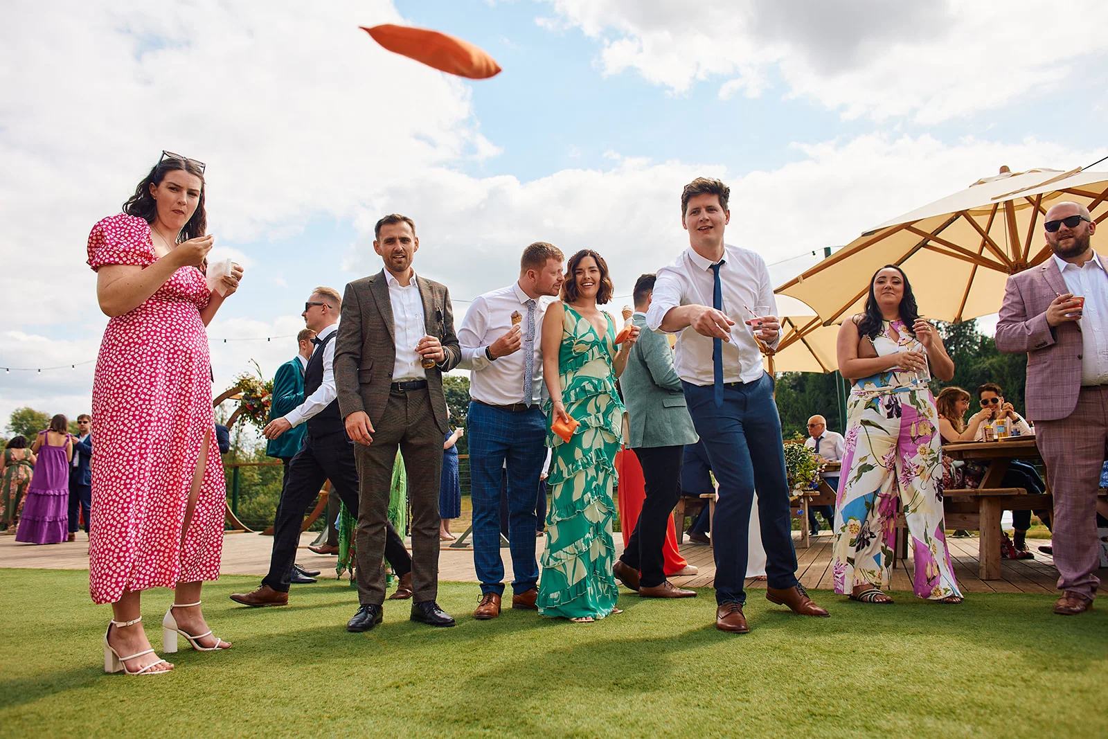 Wedding guests enjoy drinks and lawn games outdoors under a bright blue sky on a sunny day.