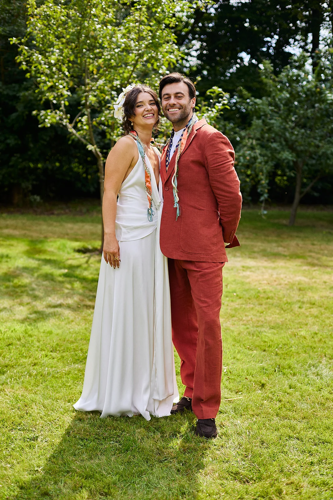 A smiling bride in a white V-neck dress and groom in a rust-coloured suit pose happily together on a lawn surrounded by trees.