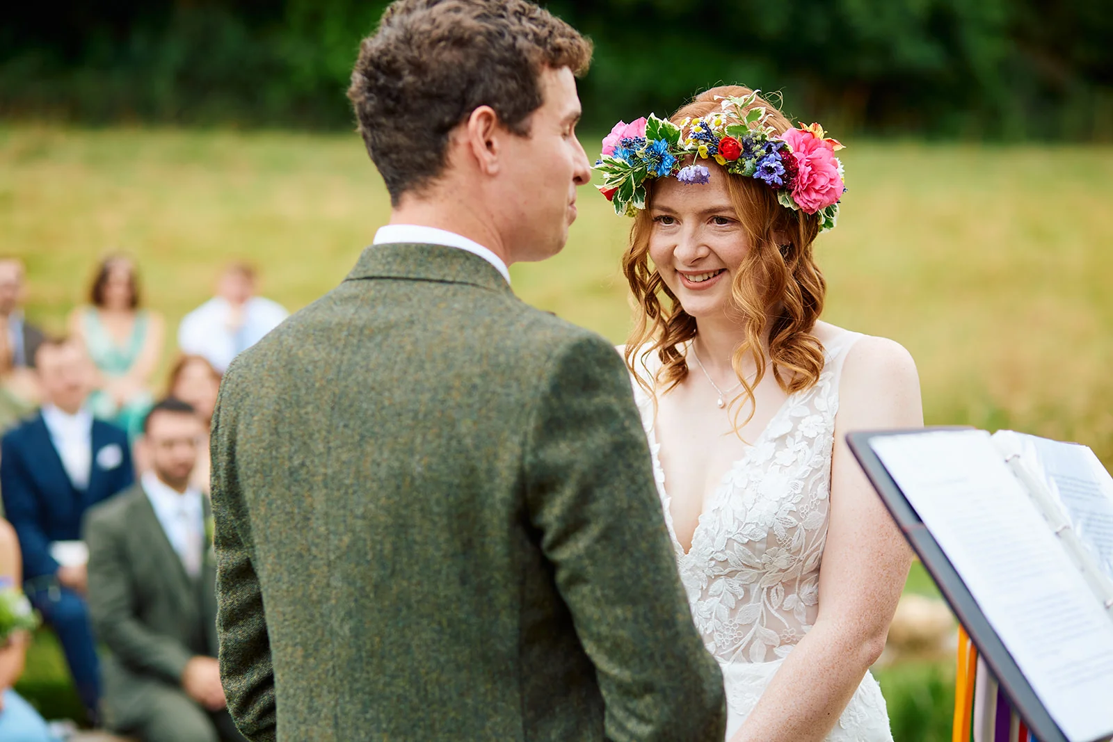 A smiling bride wearing a vibrant floral crown and a lace wedding dress gazes lovingly at her groom during an outdoor ceremony.