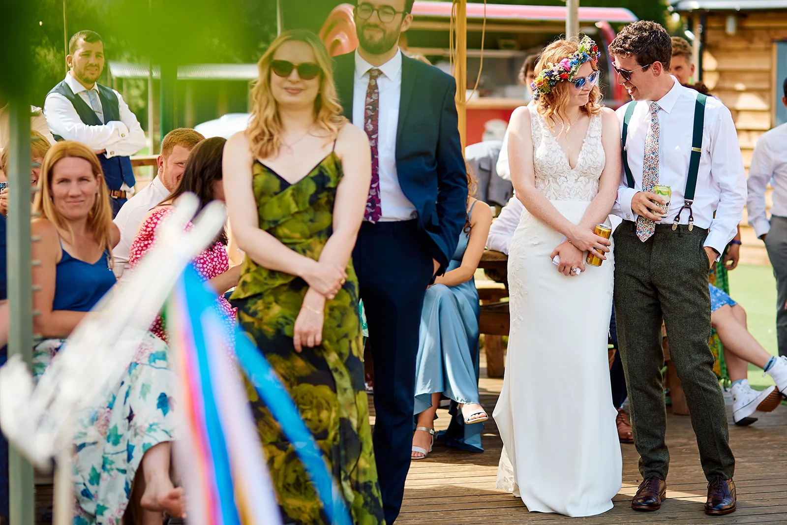 A relaxed outdoor wedding reception in the sunshine, featuring guests and the newlyweds mingling on a wooden deck while holding drinks.