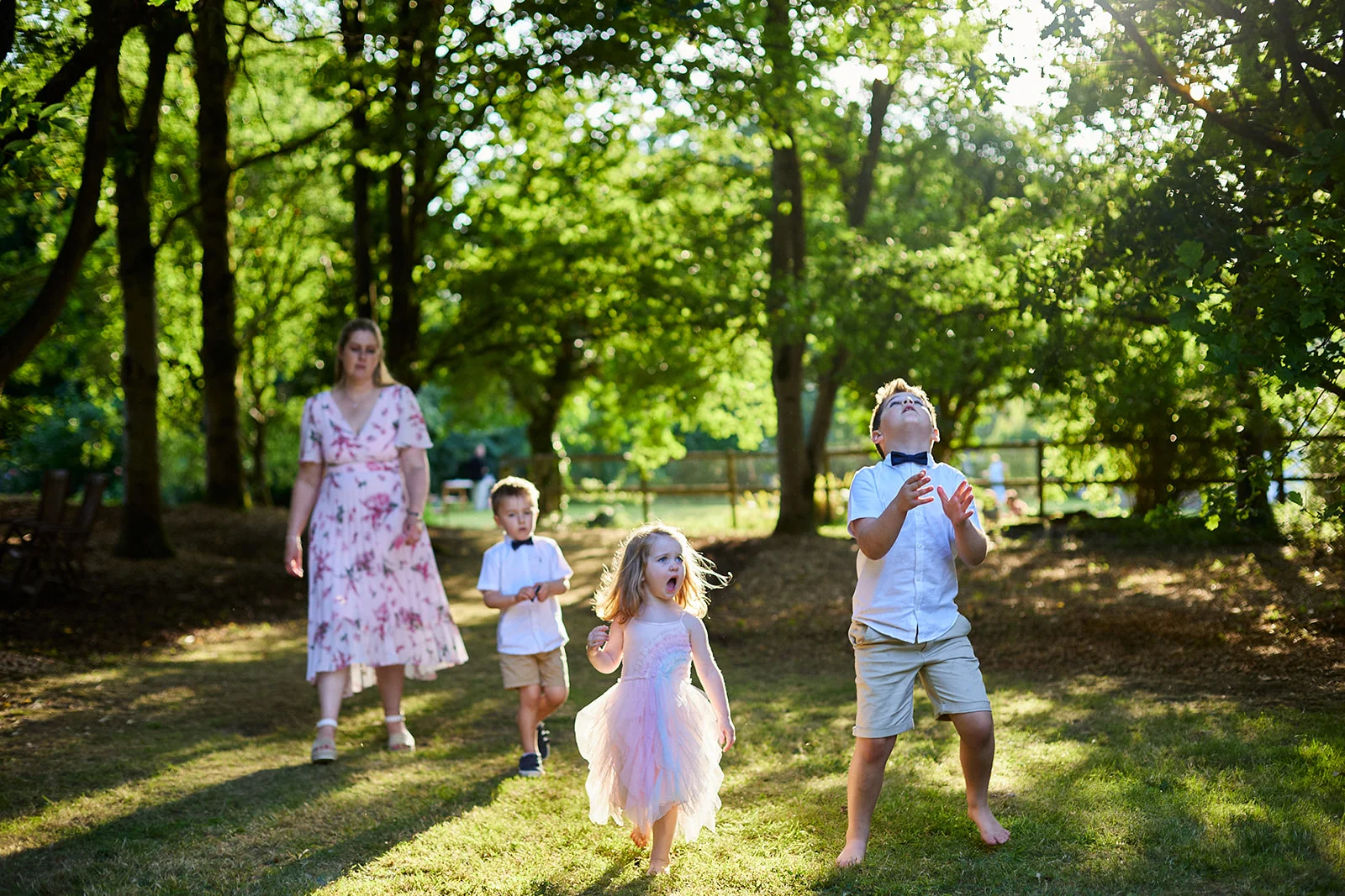 A young wedding guest in a pink dress reacts excitedly while playing outdoors in a sunlit woodland setting with other children and an adult.