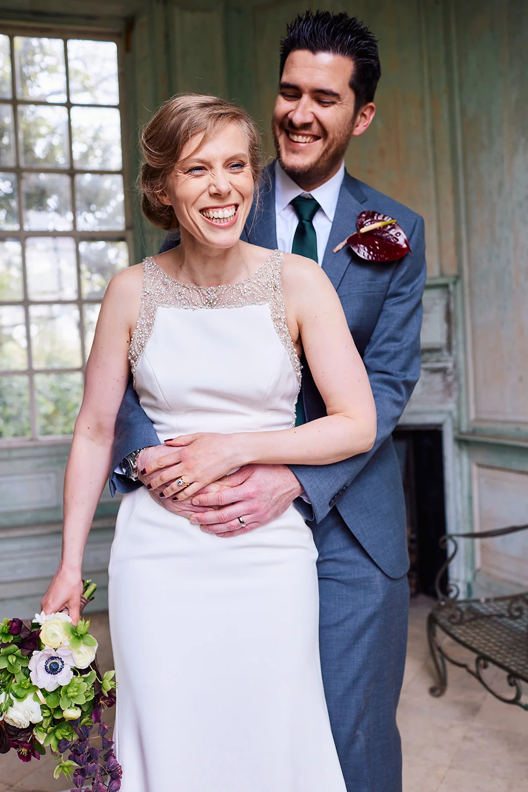 A joyful newlywed couple embrace and laugh together inside a historic manor house featuring panelled walls and large windows.
