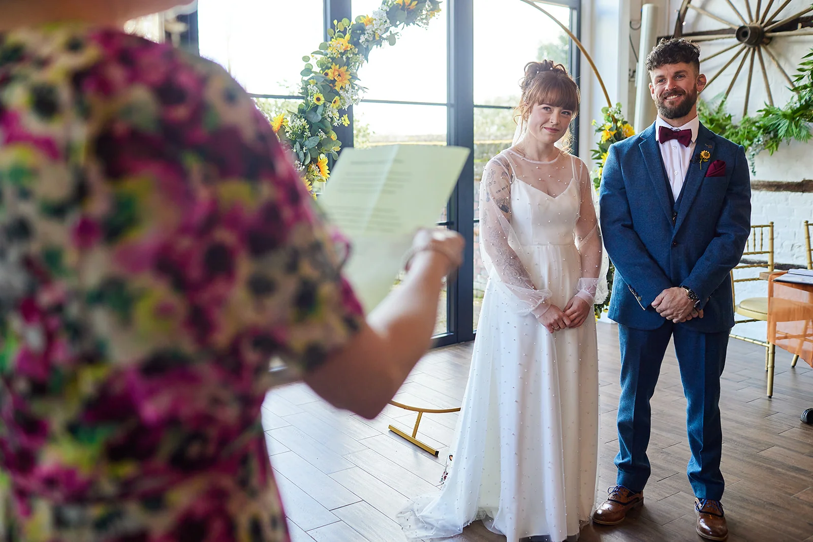 A celebrant reads vows during a wedding ceremony while the smiling bride and groom stand before a floral arch featuring bright yellow sunflowers.