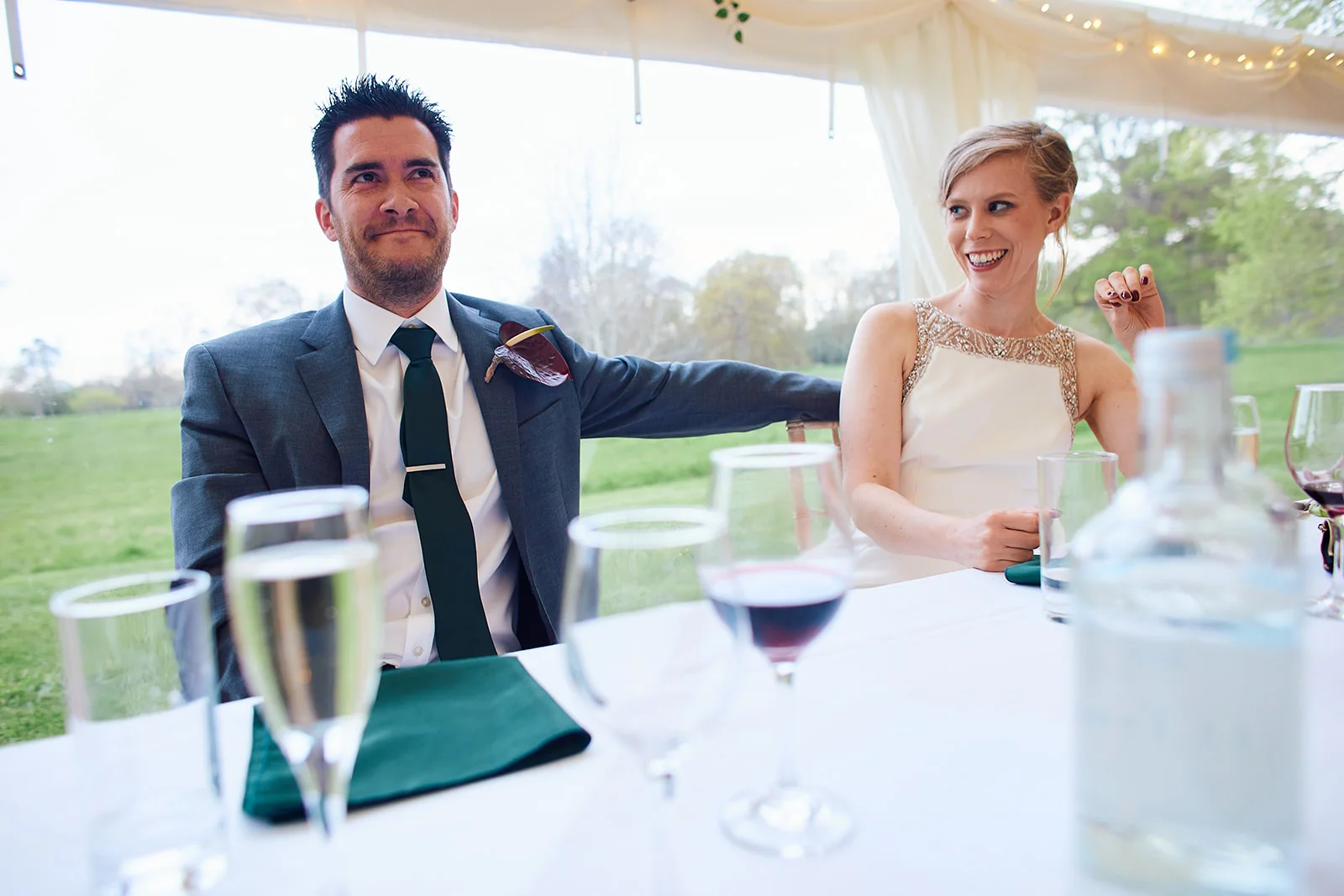 The bride and groom sit at the top table during wedding speeches inside a marquee, laughing and enjoying the moment together.