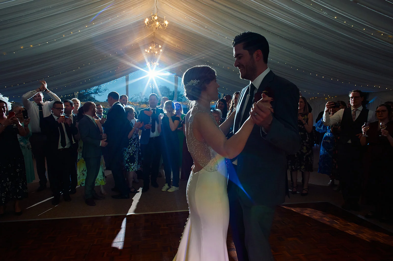 Newlyweds share their first dance surrounded by cheering guests inside a softly draped marquee illuminated by a bright sun flare from behind.