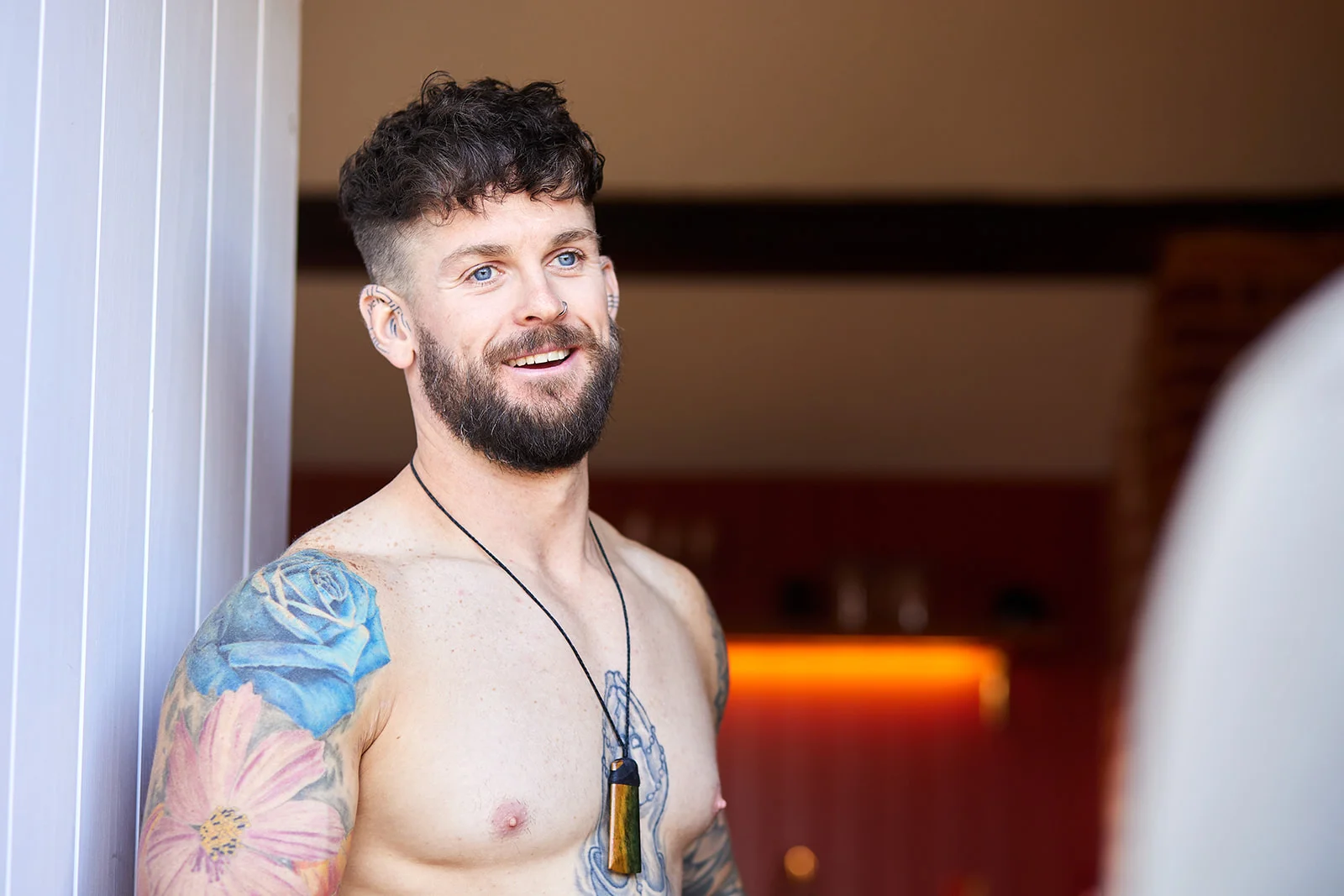 A tattooed groom smiling happily while getting ready in a bright room with natural window light.