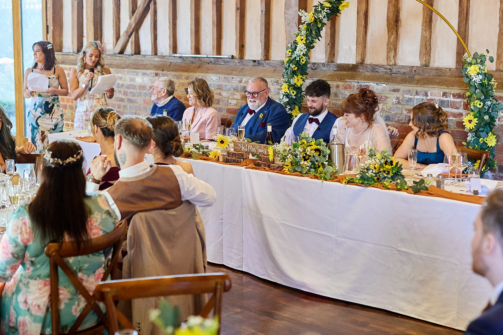 Guests enjoy speeches at a rustic barn wedding reception, seated at a long top table decorated with sunflowers and foliage.
