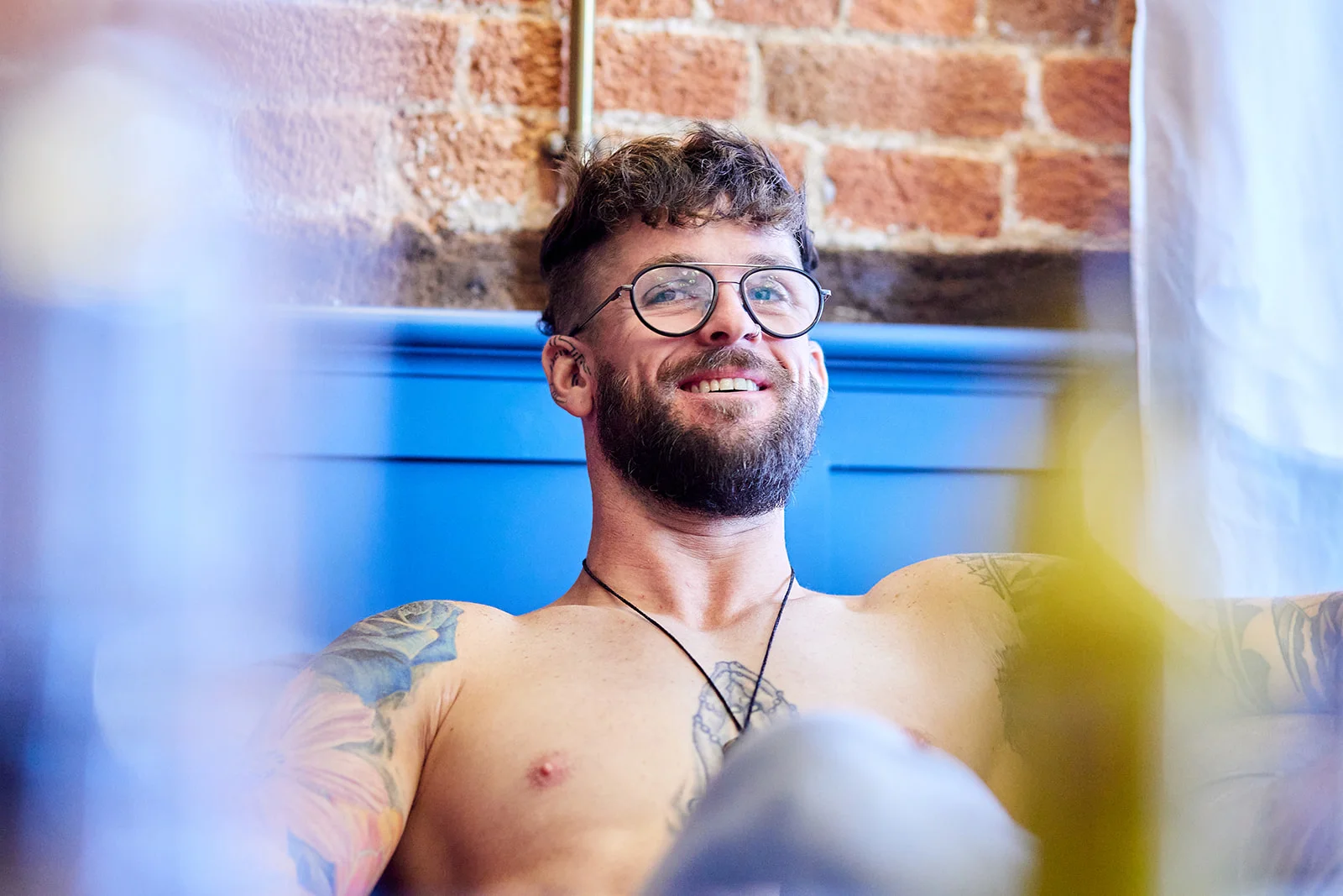 A tattooed groom smiles happily while getting ready in a room with an exposed brick wall.