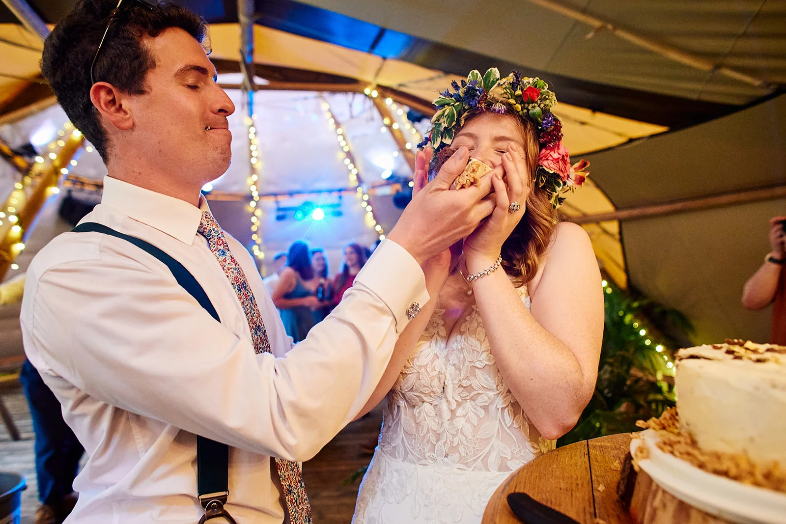 A groom playfully feeds cake to his laughing bride inside a marquee decorated with fairy lights.