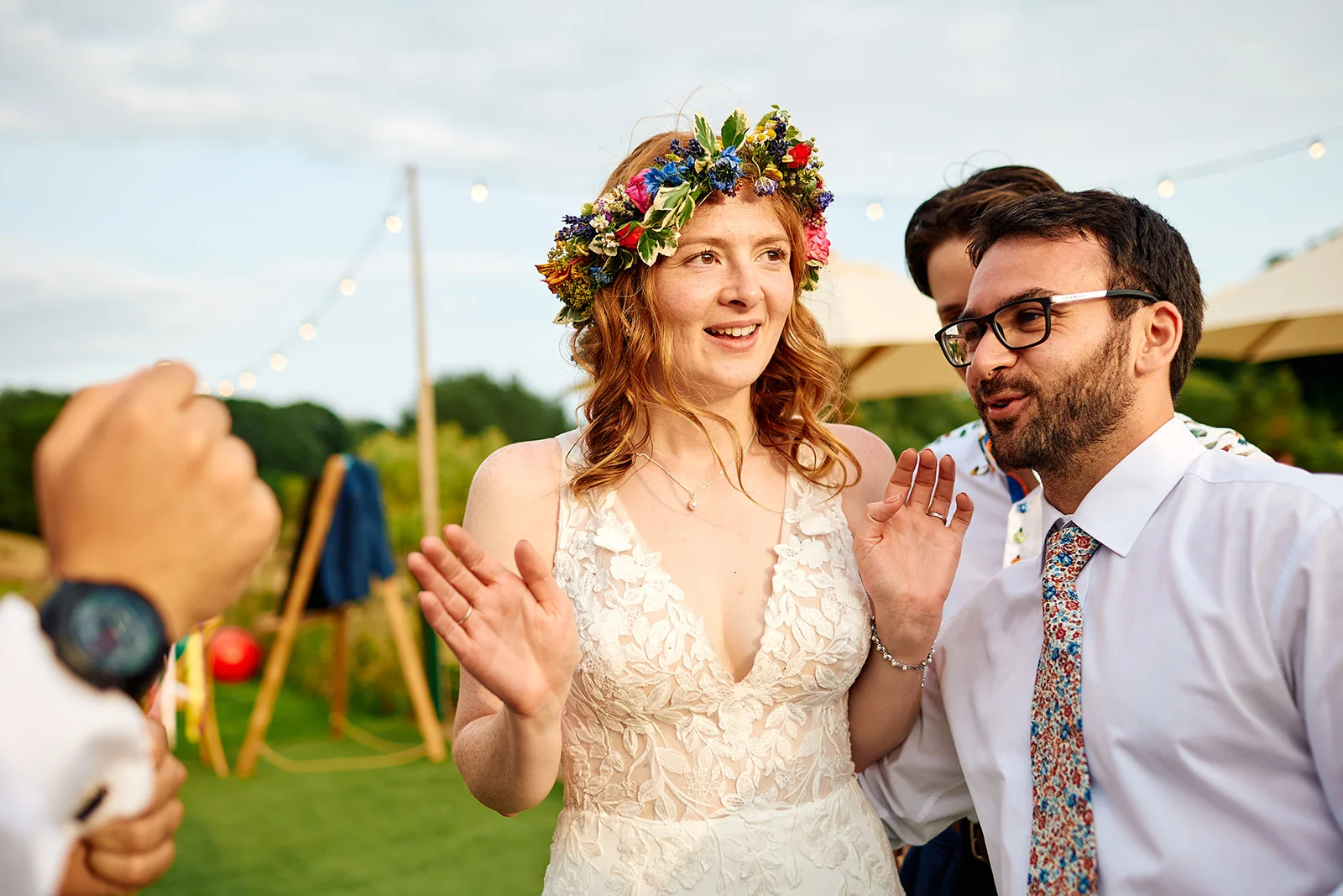 A joyful outdoor wedding scene featuring a laughing bride wearing a vibrant fresh flower crown and a lace gown alongside her groom in an open-collared shirt and patterned tie.
