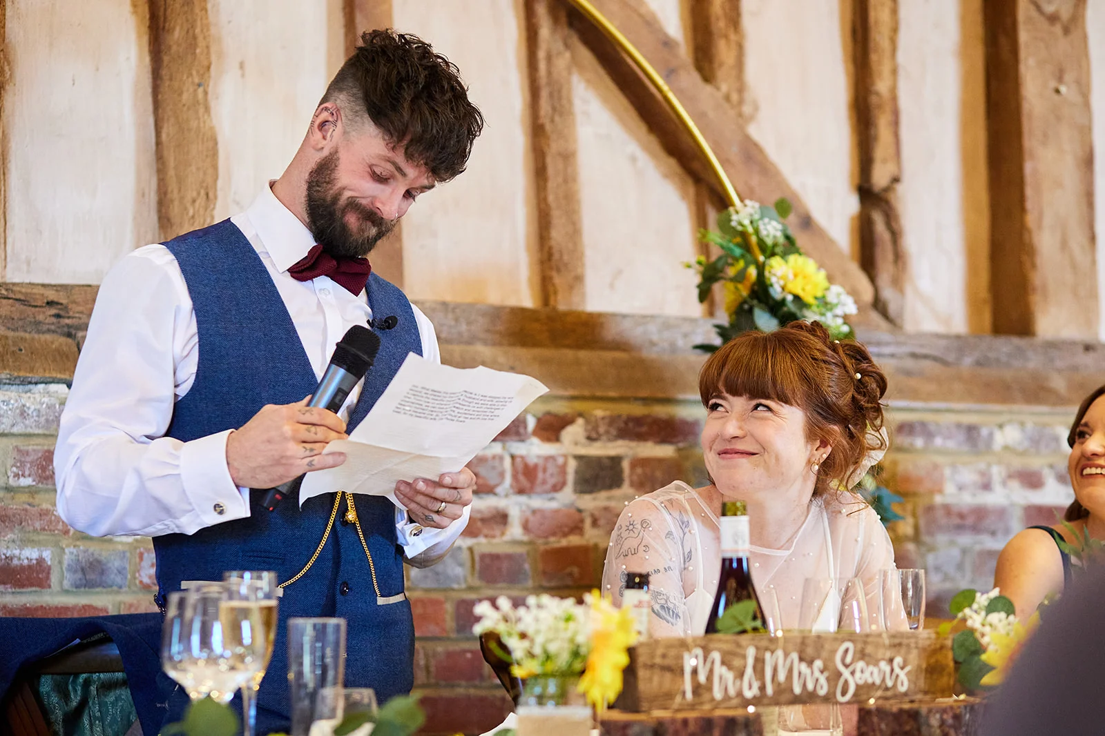 A groom delivers an emotional speech while reading from paper, holding a microphone in a rustic barn venue with exposed brick walls, as his smiling bride looks up at him adoringly from the top table.