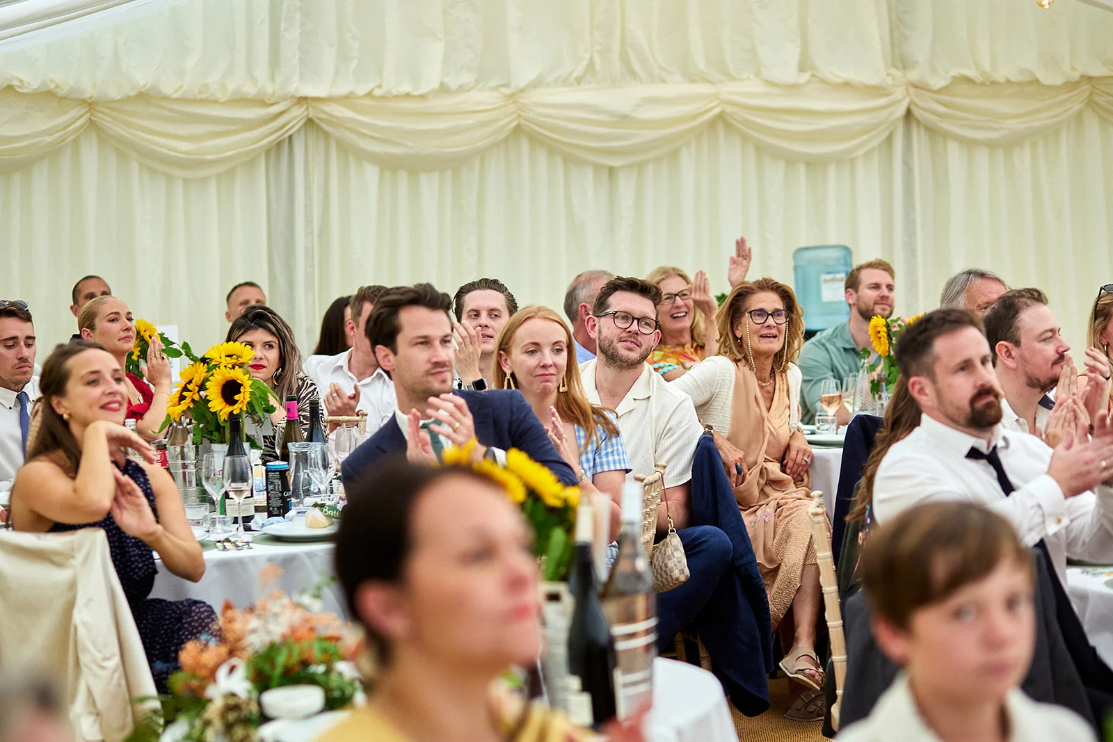 Wedding guests sit inside a draped marquee, laughing and clapping enthusiastically during the speeches.