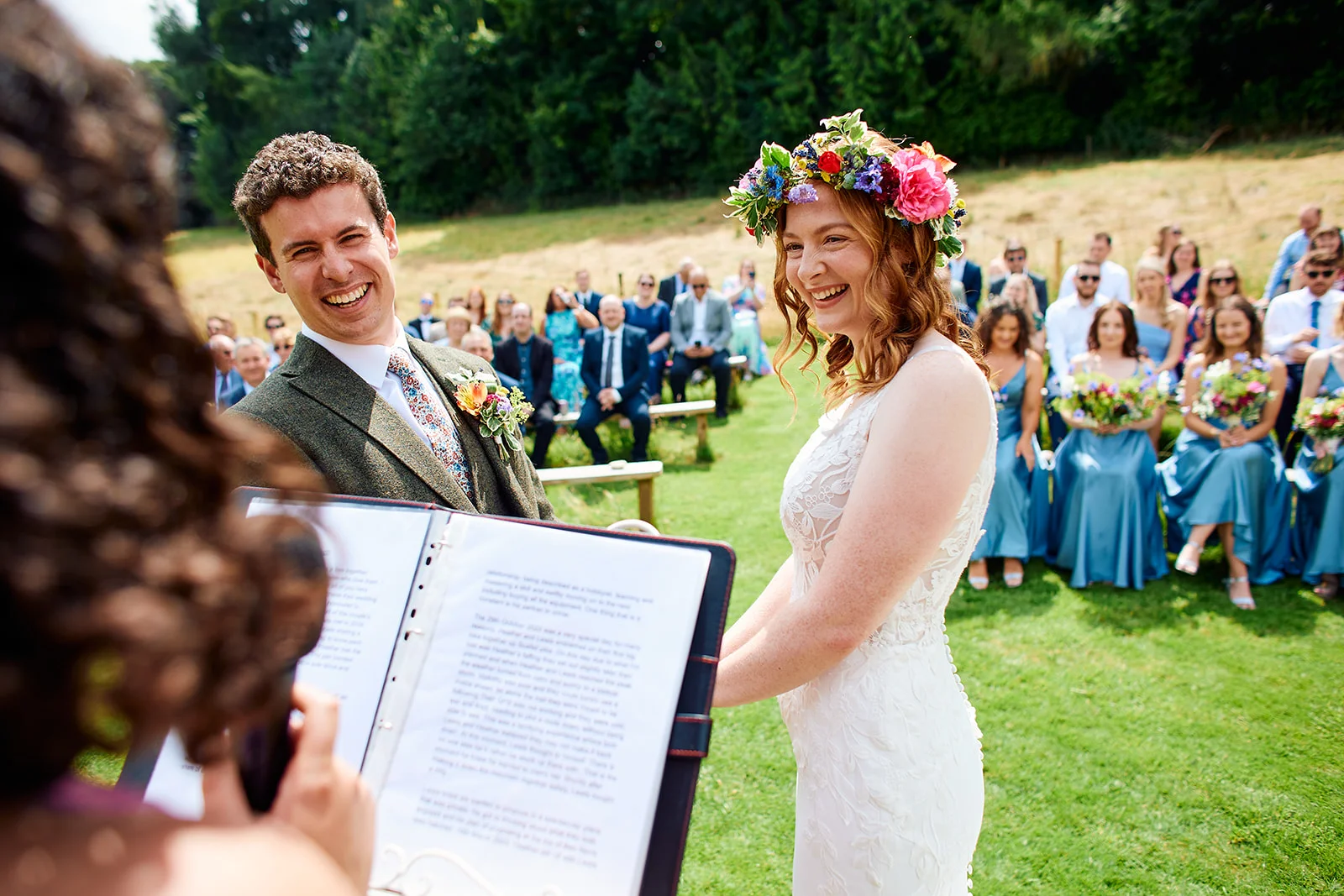 A smiling bride wearing a vibrant floral crown and a lace wedding dress stands happily with the groom at an outdoor humanist ceremony on a sunny day.