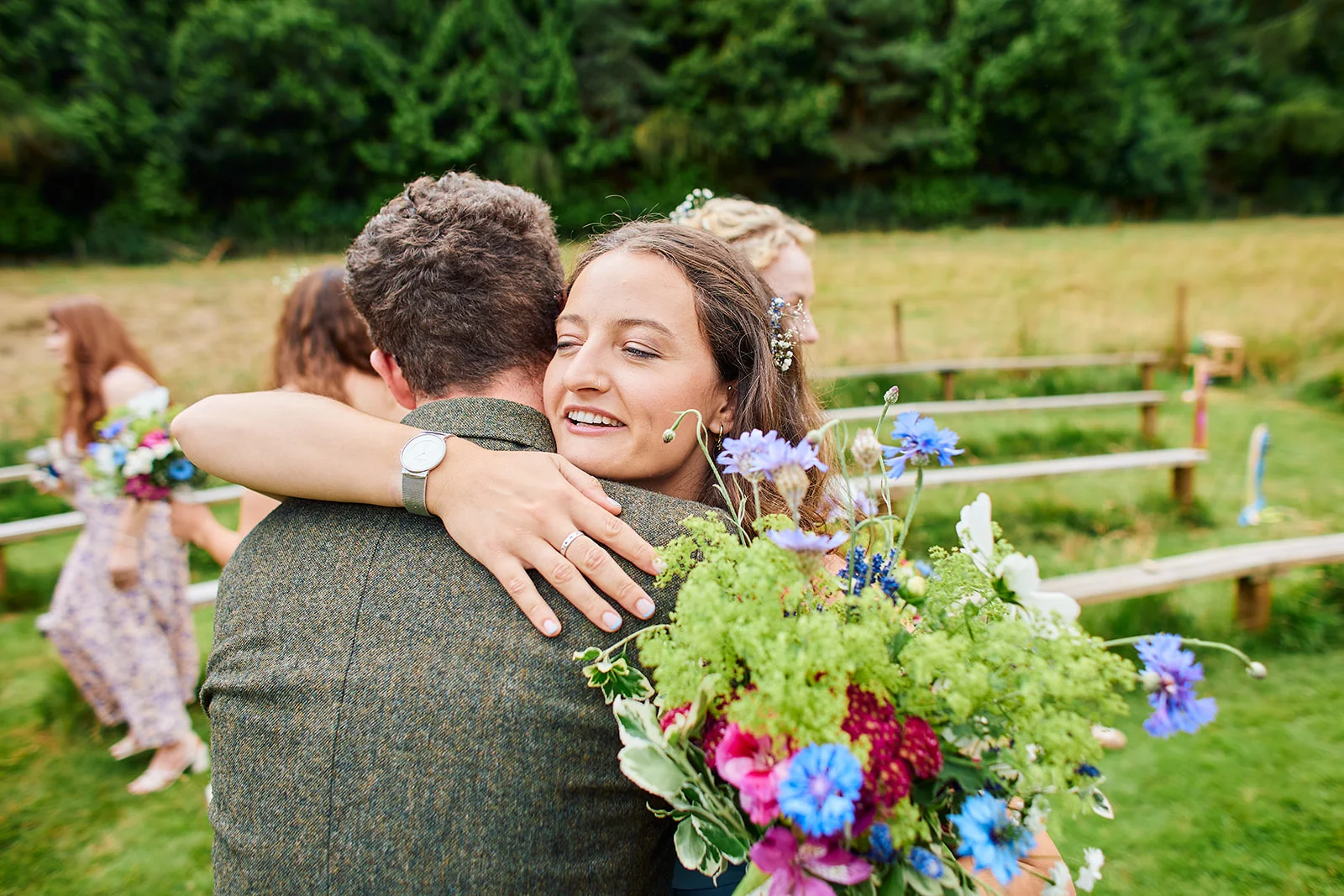 A smiling bride wearing her engagement ring embraces the groom in a tweed jacket while holding a colourful wildflower bouquet outdoors.