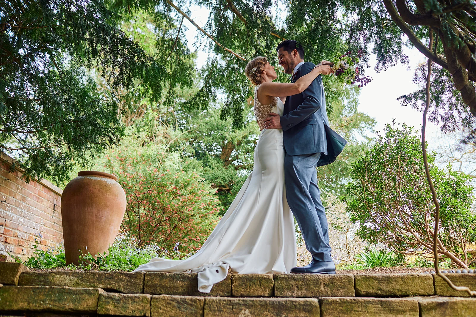 A newlywed couple embraces lovingly nose-to-nose on a stone terrace framed by lush green trees and garden foliage.
