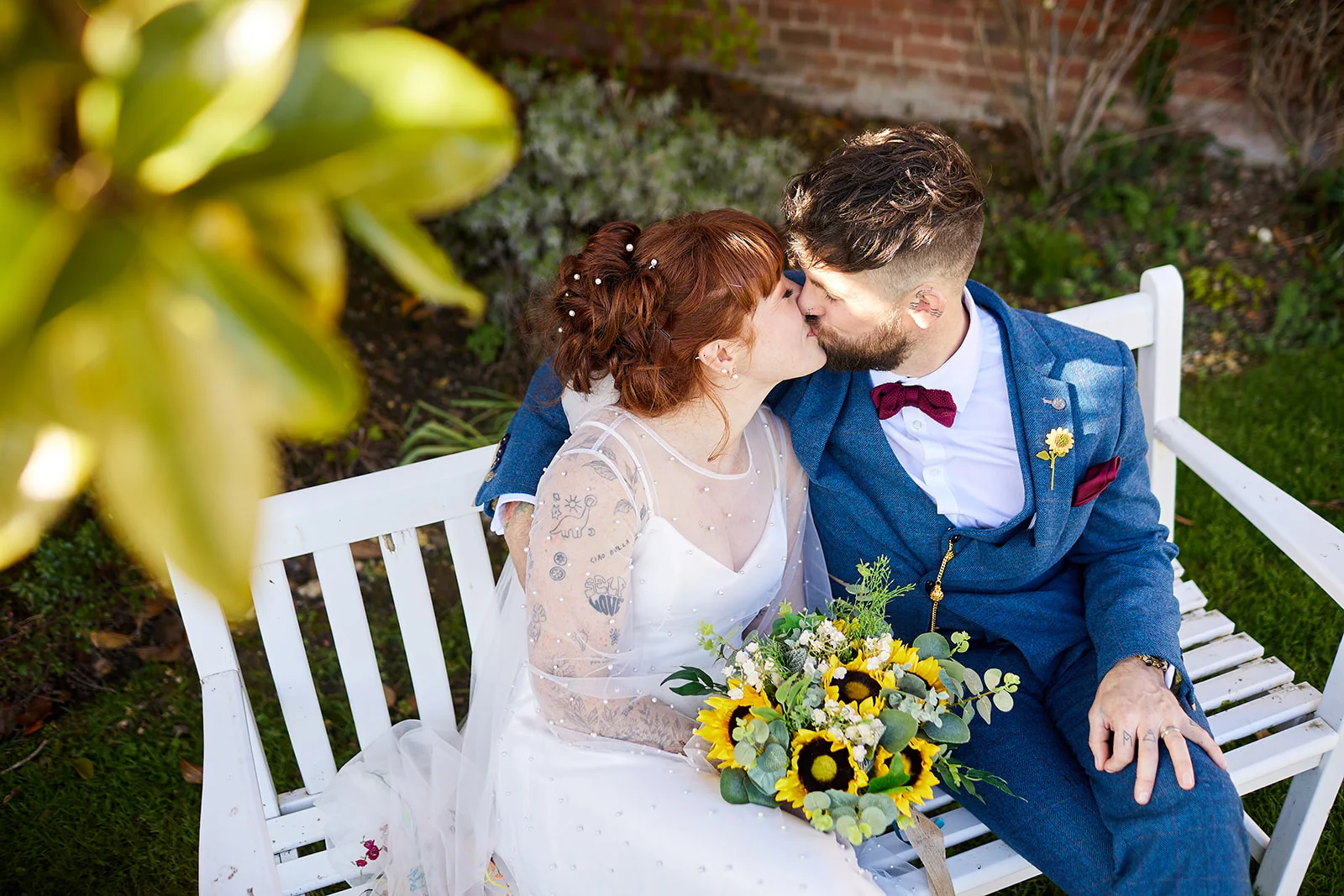 A newlywed couple shares a tender kiss while seated together on a white wooden garden bench outdoors.