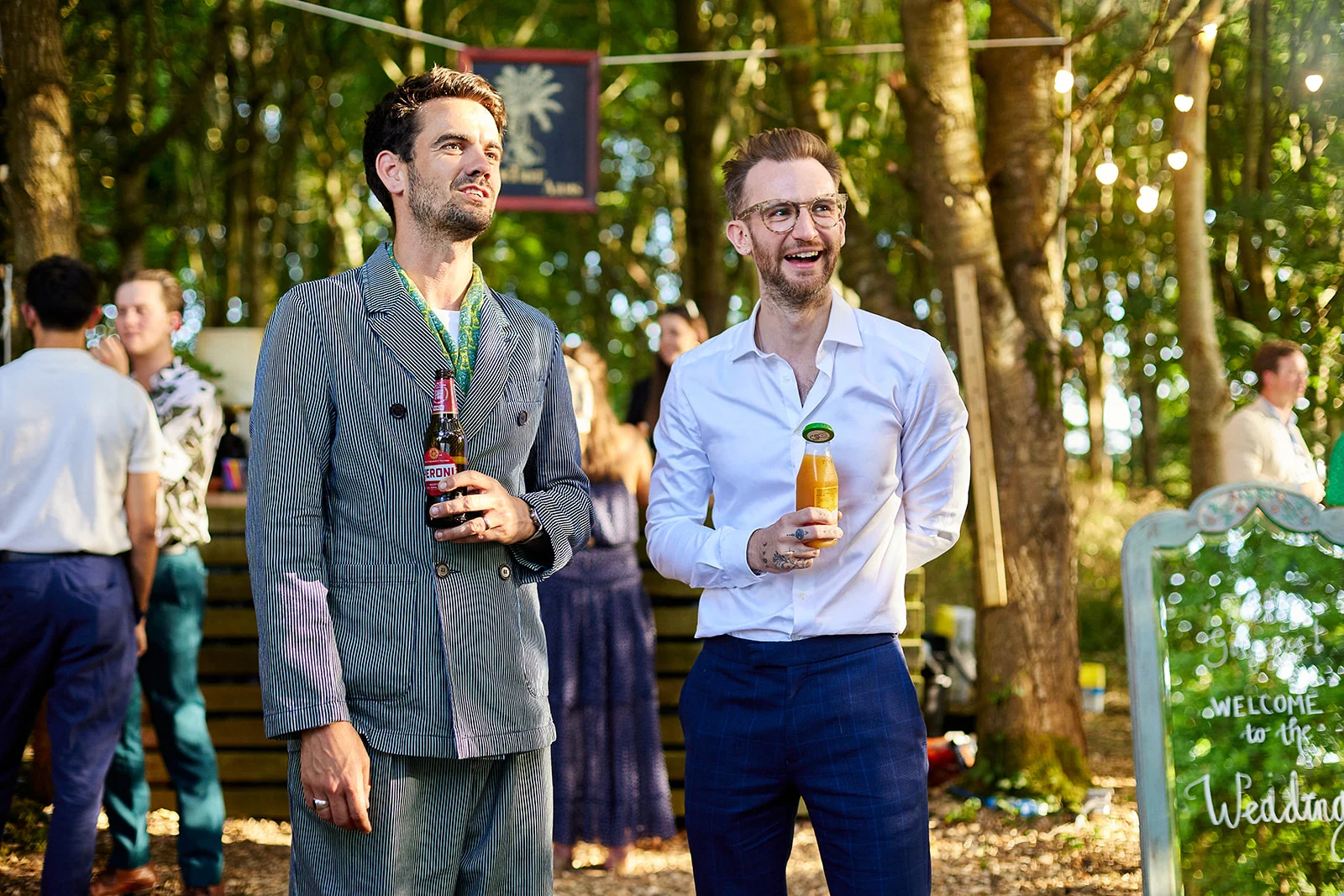Two smiling grooms share a joyful moment outdoors at a woodland wedding drinks reception, surrounded by trees and dappled sunlight.