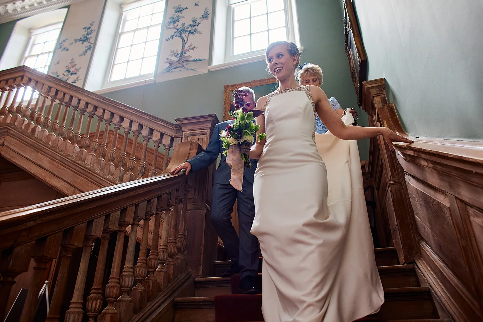 A smiling bride holding a bouquet descends a grand wooden staircase alongside her father inside a historic country house.