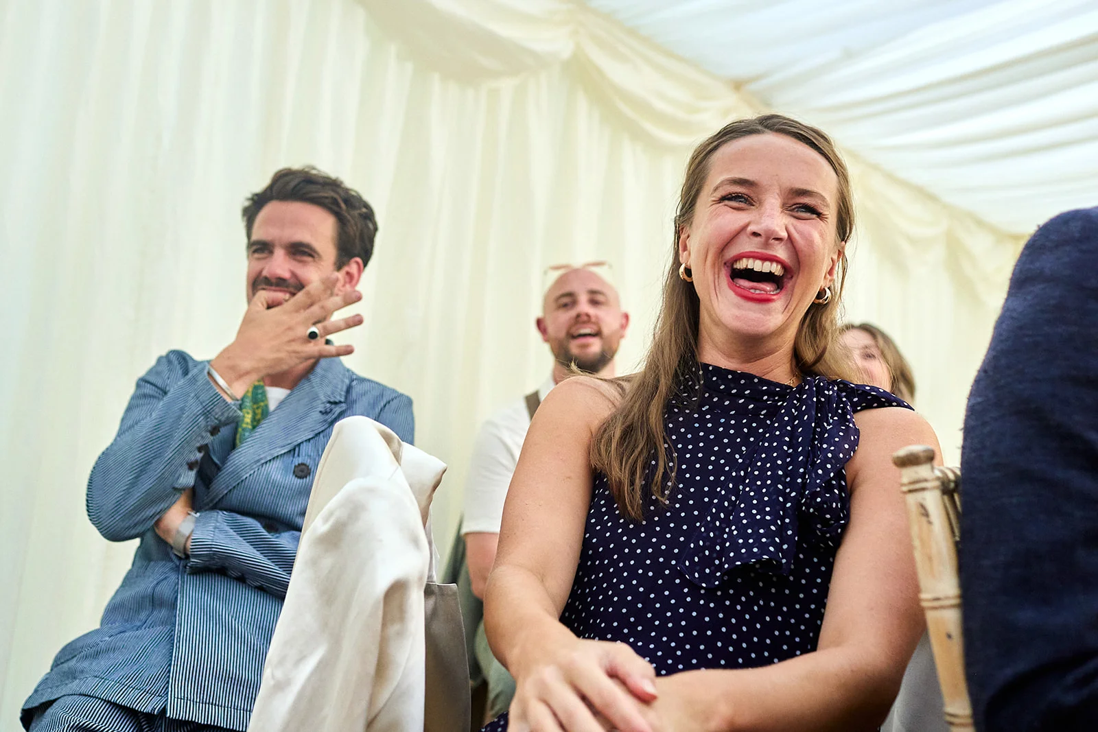 A smiling wedding guest in a polka-dot dress laughs heartily while seated among guests inside a draped marquee.