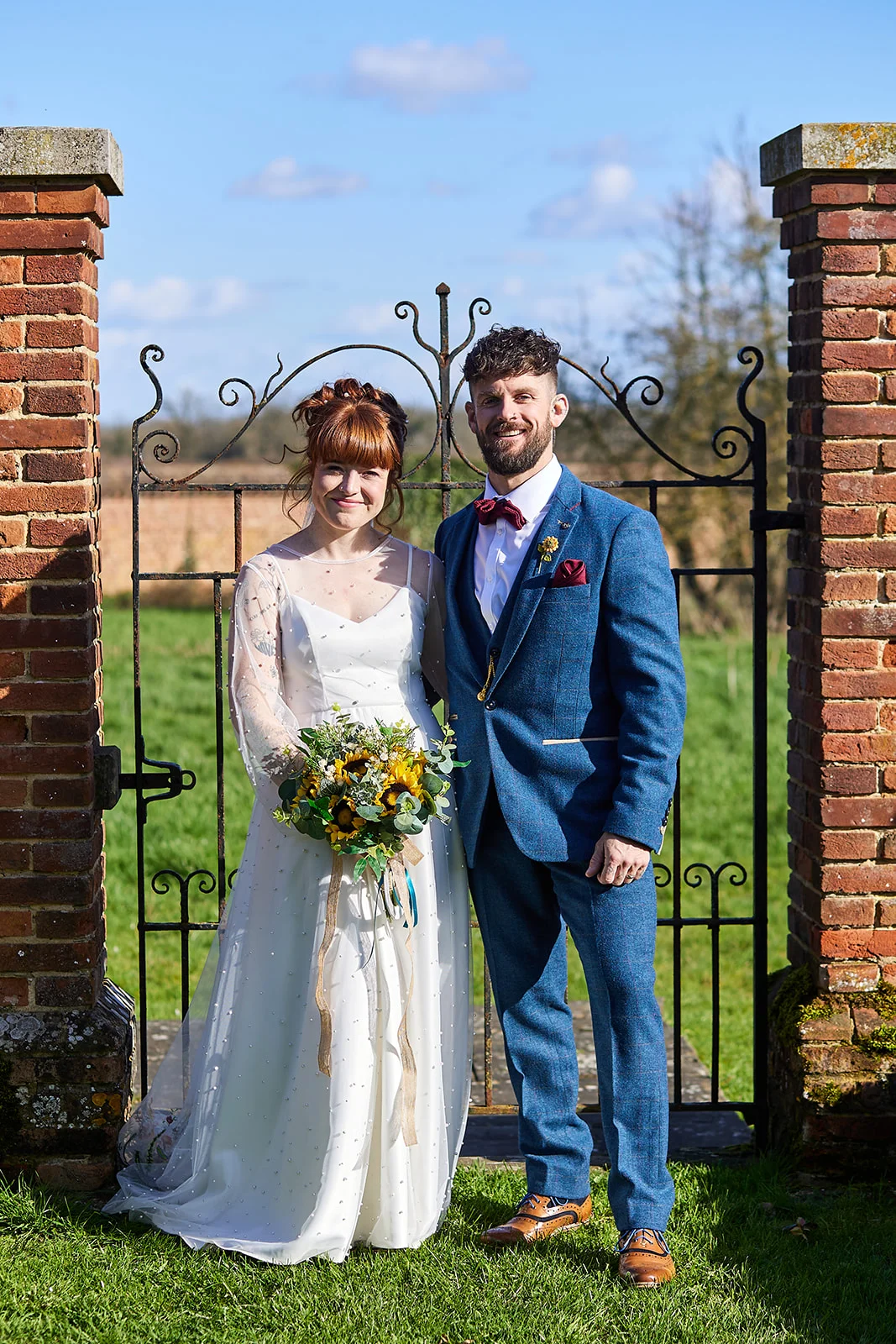 A newlywed couple stands smiling outside in front of an iron gate set between brick pillars, with the bride holding a vibrant sunflower bouquet and wearing a pearl-embellished gown alongside the groom in a sharp blue tweed suit.