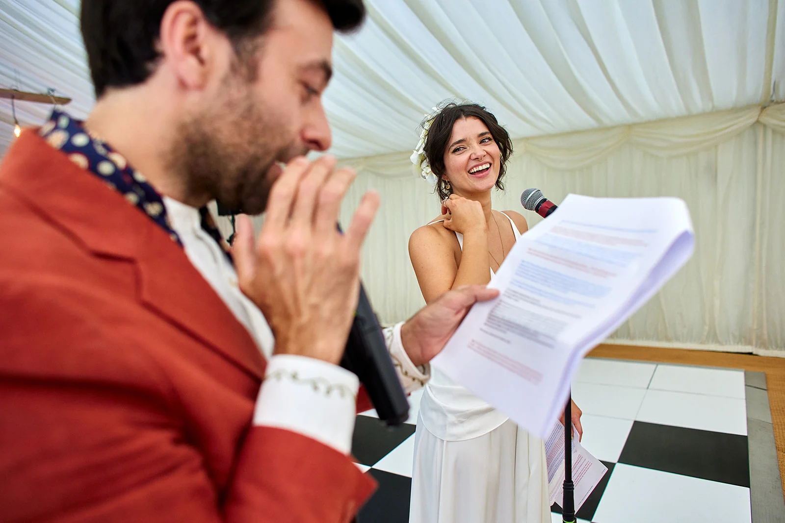 A groom speaks into a microphone while reading notes, with the bride laughing joyfully in the background inside a marquee reception.