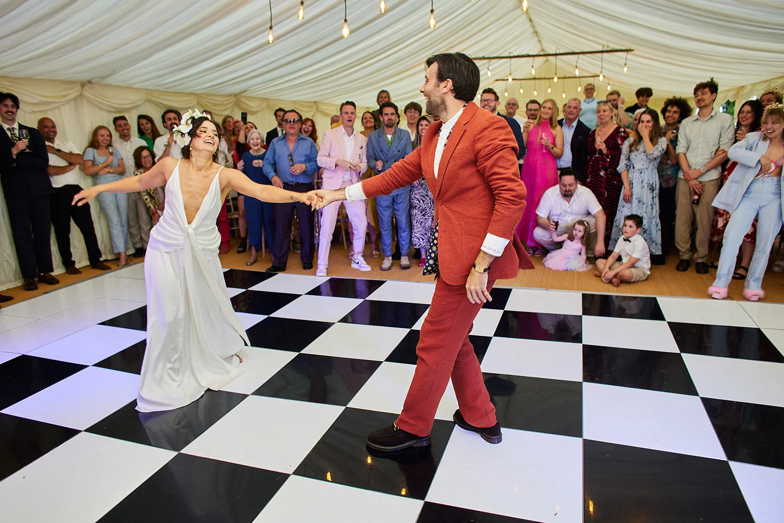 The newlyweds share a joyful first dance on a black and white checkered dance floor inside a marquee surrounded by cheering wedding guests.