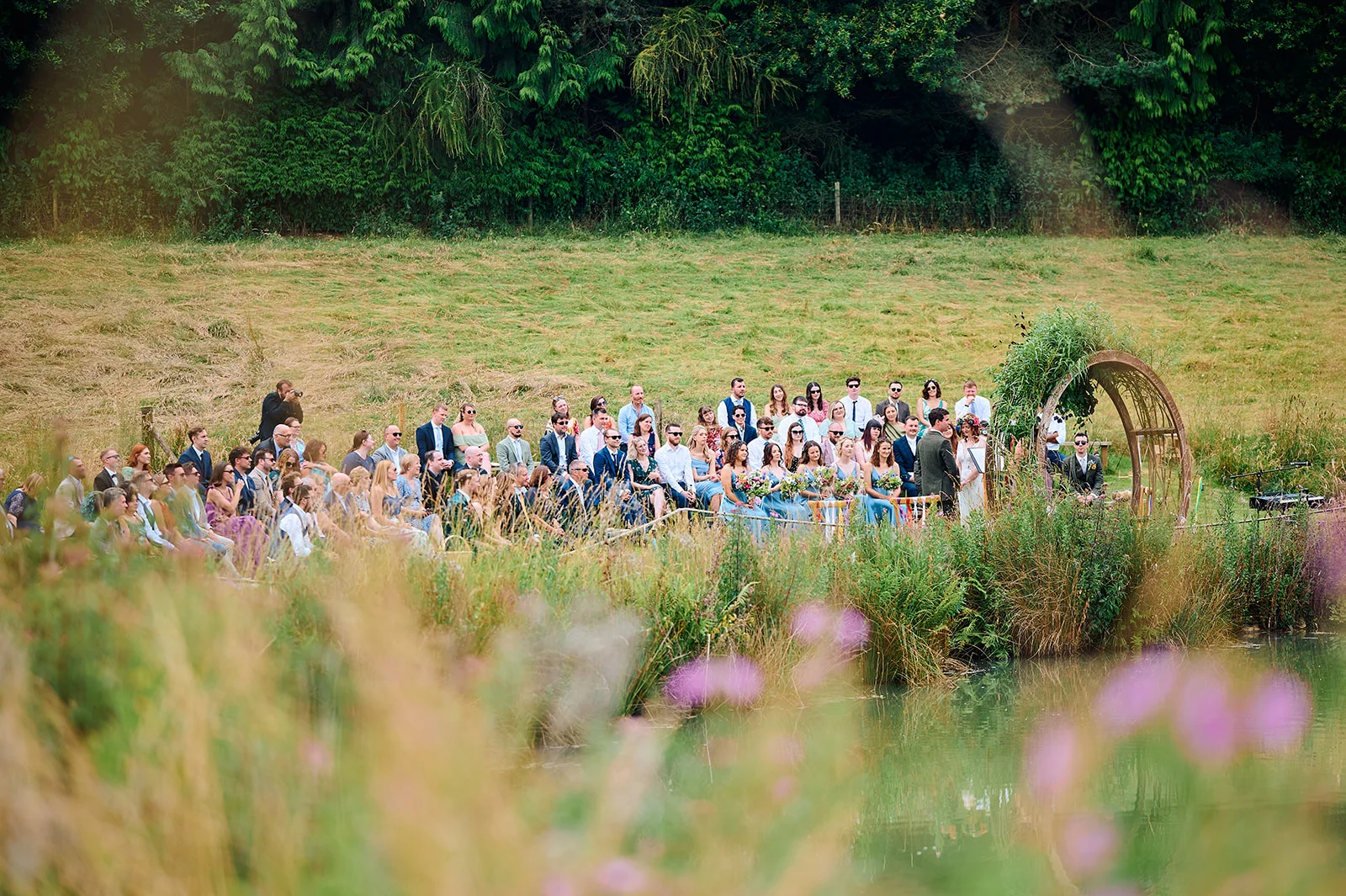 An outdoor wedding ceremony takes place by a serene lake with guests seated on a grassy bank beneath a circular floral arch.