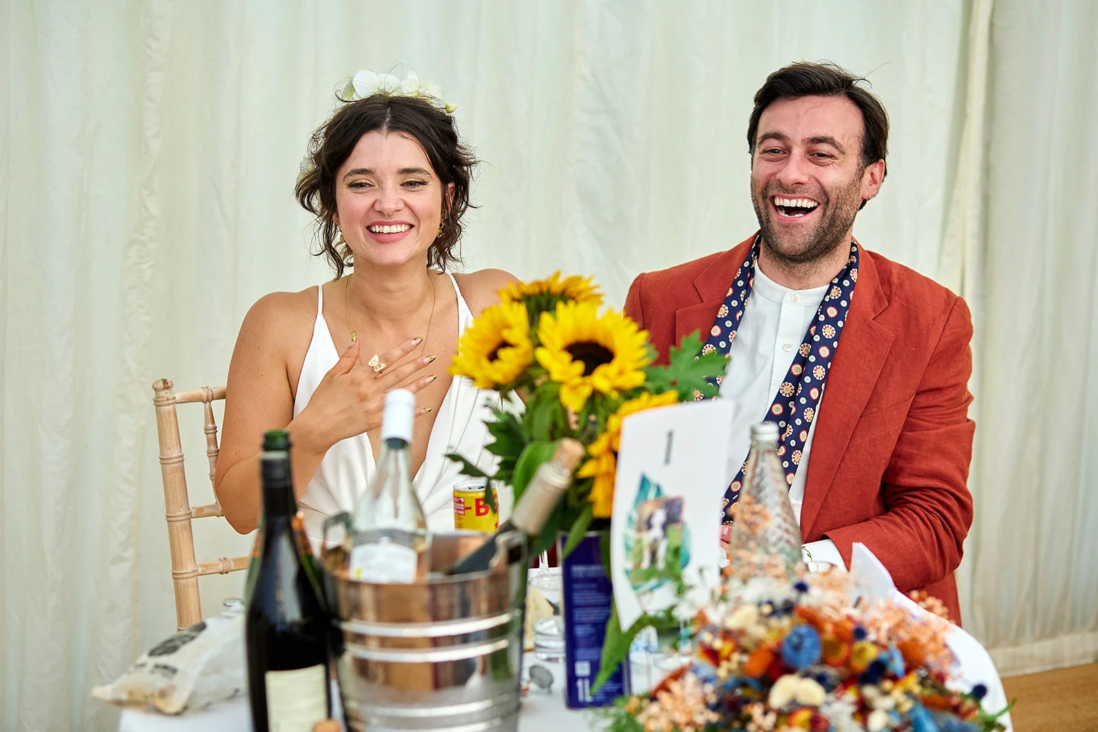 A newlywed couple sitting together at the top table inside a marquee, laughing heartily during wedding speeches with sunflowers on the table.