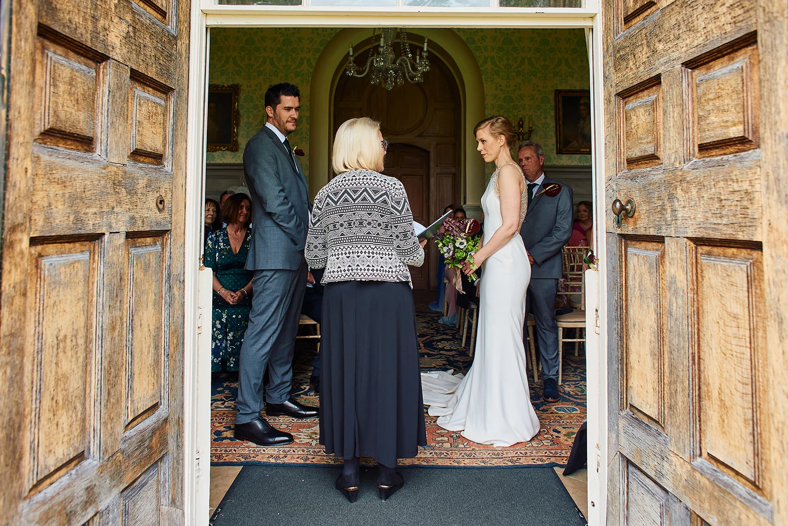 A bride in a sleek column dress and groom in a grey suit exchange vows during a civil ceremony framed by large wooden doors.