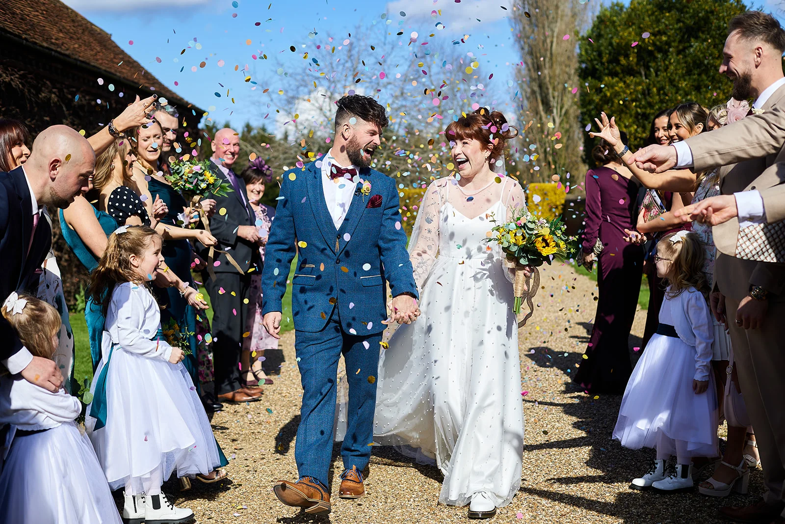 A joyful newlywed couple walks hand in hand down a gravel path while cheering guests shower them in colourful confetti on a sunny day.