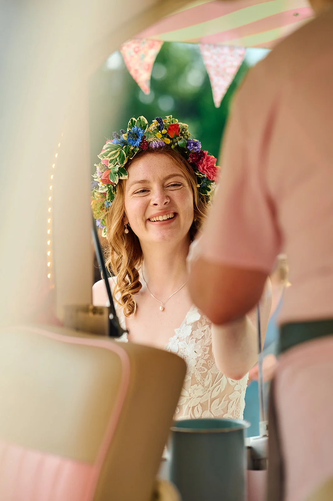 A smiling bride wearing a colourful fresh flower crown and lace wedding dress is captured through a window during an outdoor celebration.
