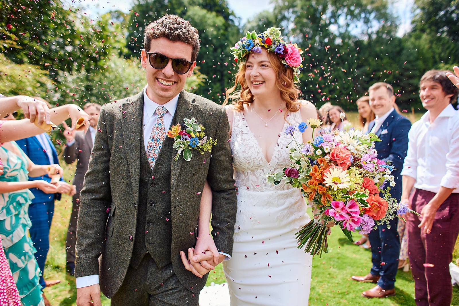 A joyful newlywed couple walks hand in hand through a shower of colourful confetti, smiling widely as wedding guests cheer them on in a lush green garden.