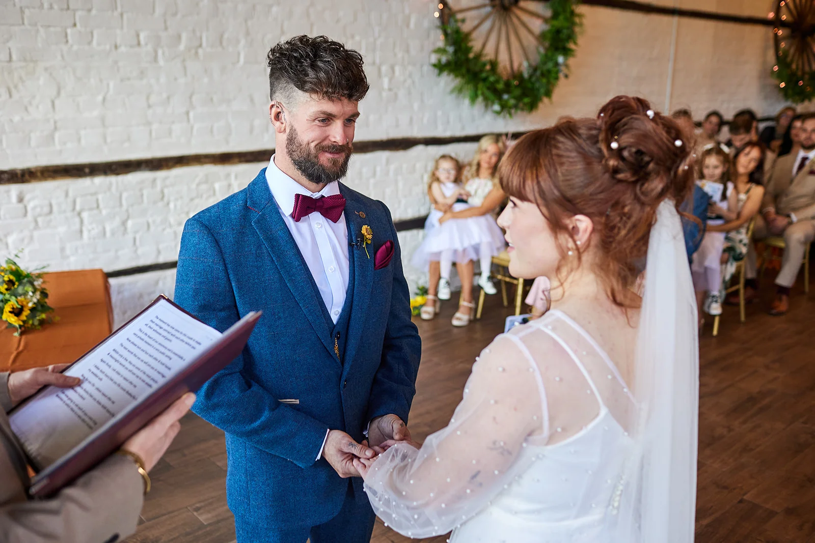 A bride and groom exchange vows during their indoor civil ceremony, with the groom wearing a blue tweed suit and the bride in a long-sleeved gown adorned with pearls.