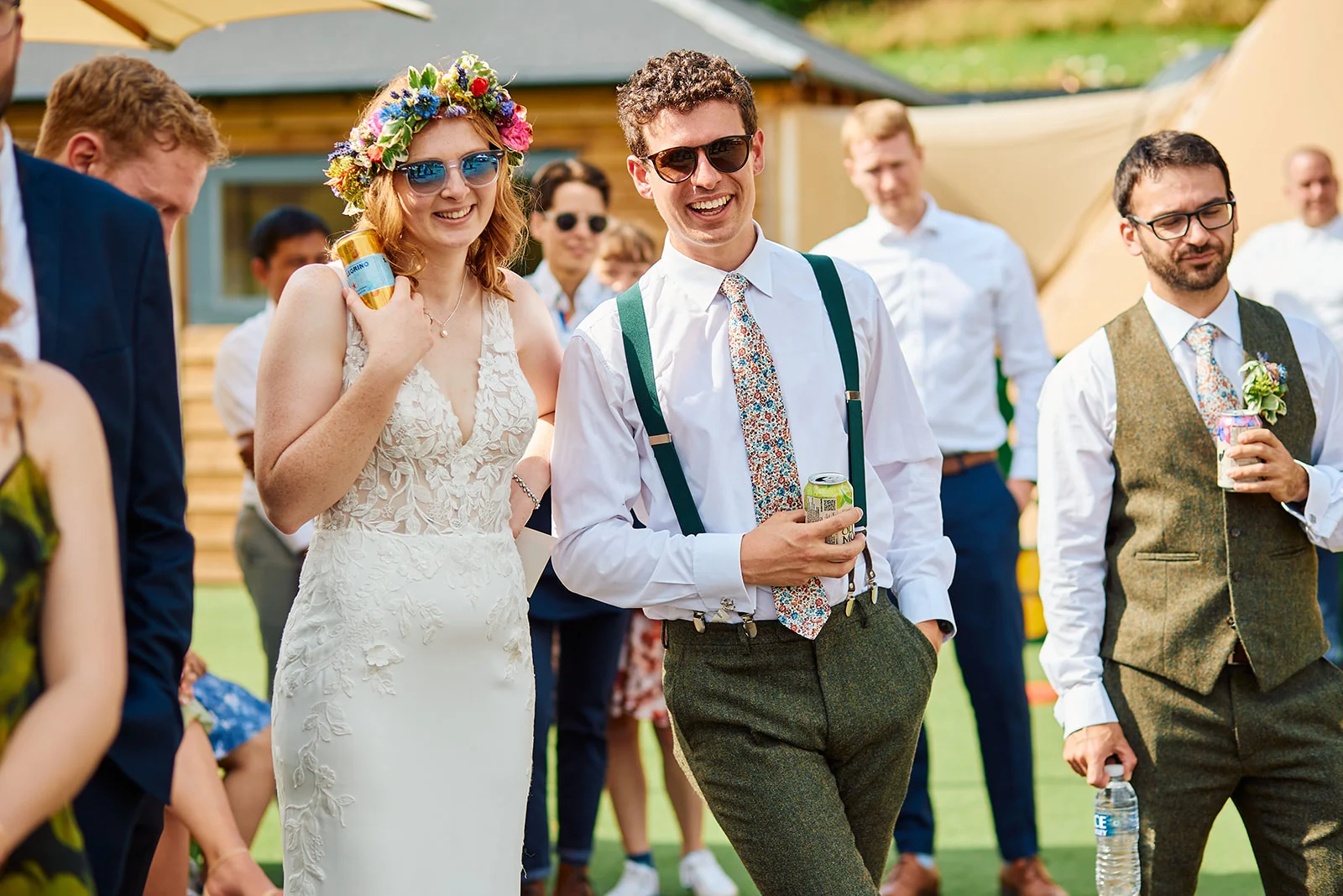 A smiling bride wearing a flower crown and sunglasses stands alongside her groom in tweed trousers, braces and a floral tie during an outdoor wedding celebration.