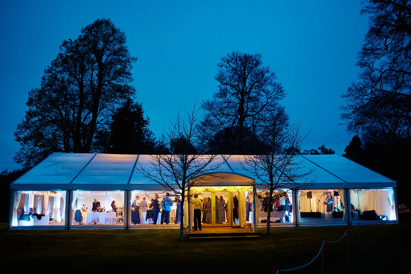 A glowing marquee wedding tent stands on a lawn at blue hour, illuminated from within while guests mingle during the evening celebration.