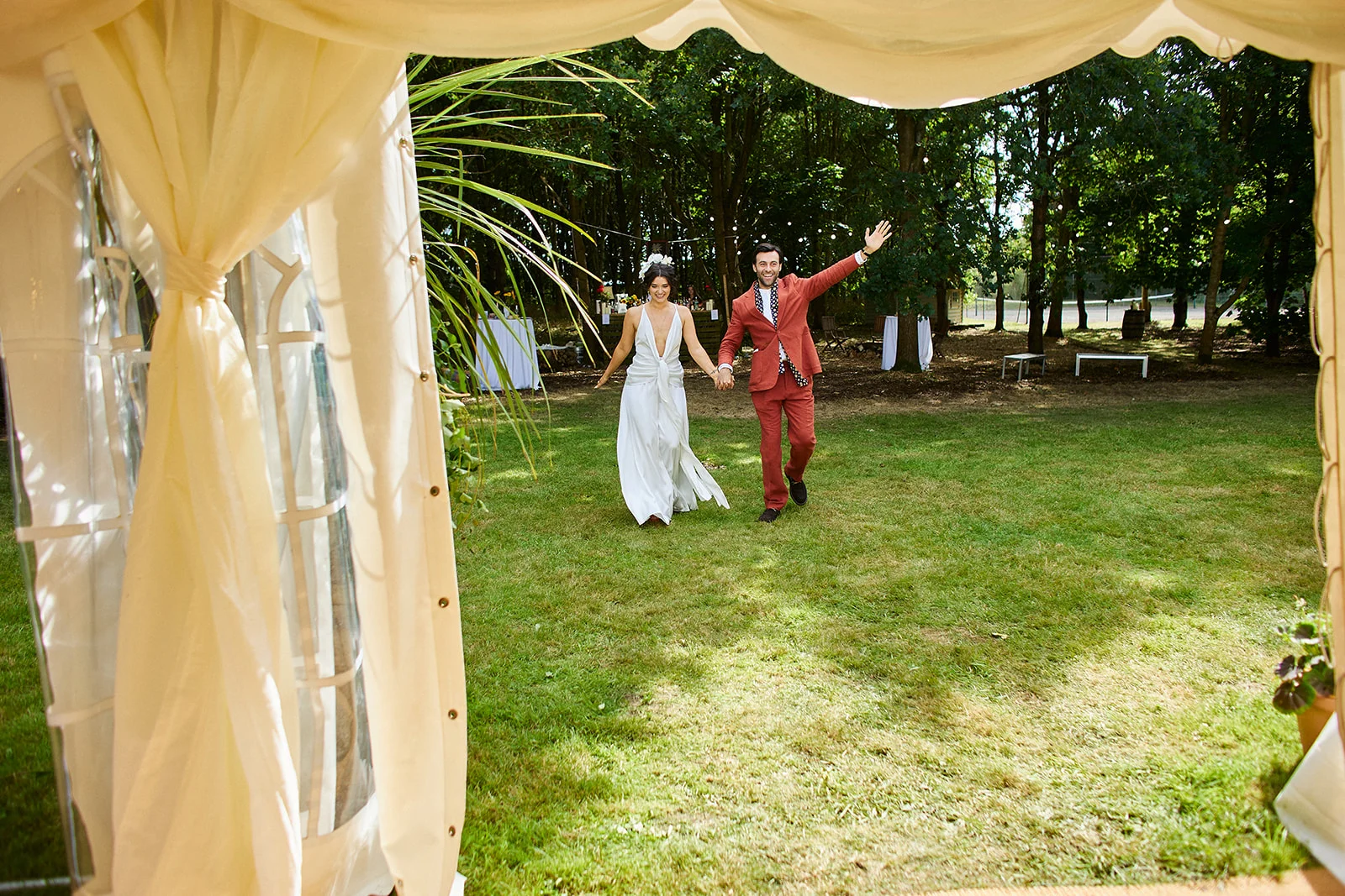 A newlywed couple run hand in hand across a grassy lawn towards a marquee, with the groom waving happily and the bride wearing a flowing white gown with a plunging neckline.