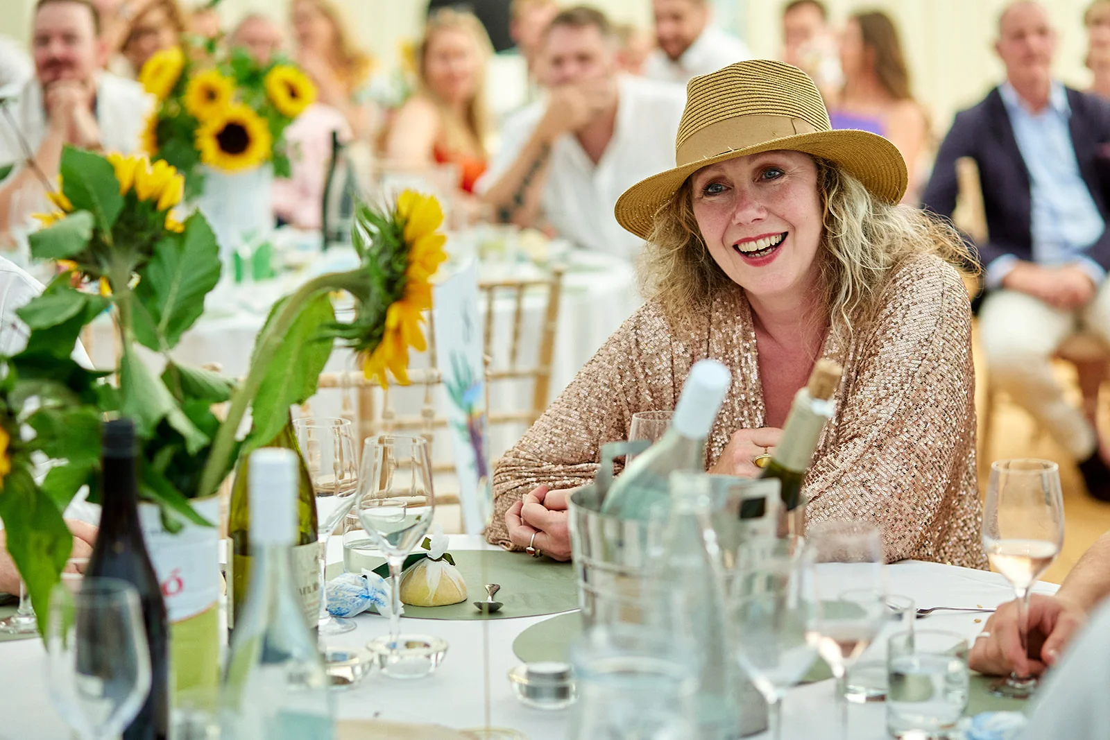 A guest wearing a stylish straw hat and a shimmering gold top laughs happily at a wedding breakfast table adorned with sunflowers.