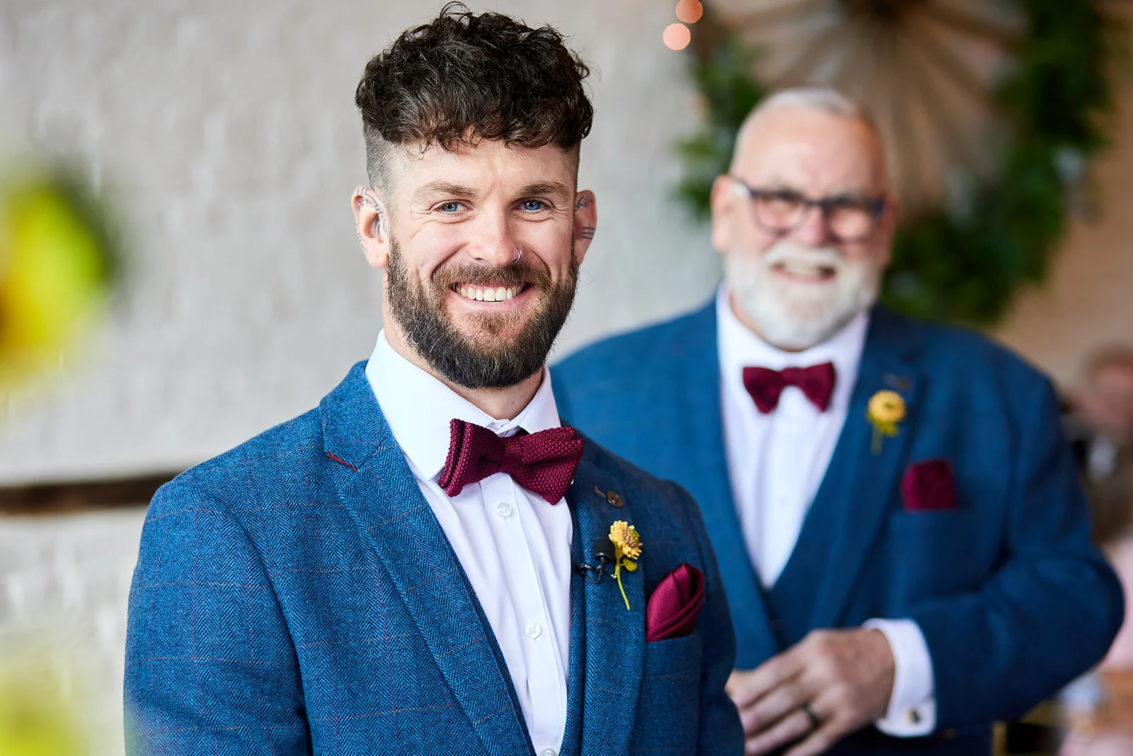 A smiling groom with a beard and patterned blue suit stands sharply in focus while his partner looks on affectionately from the background.