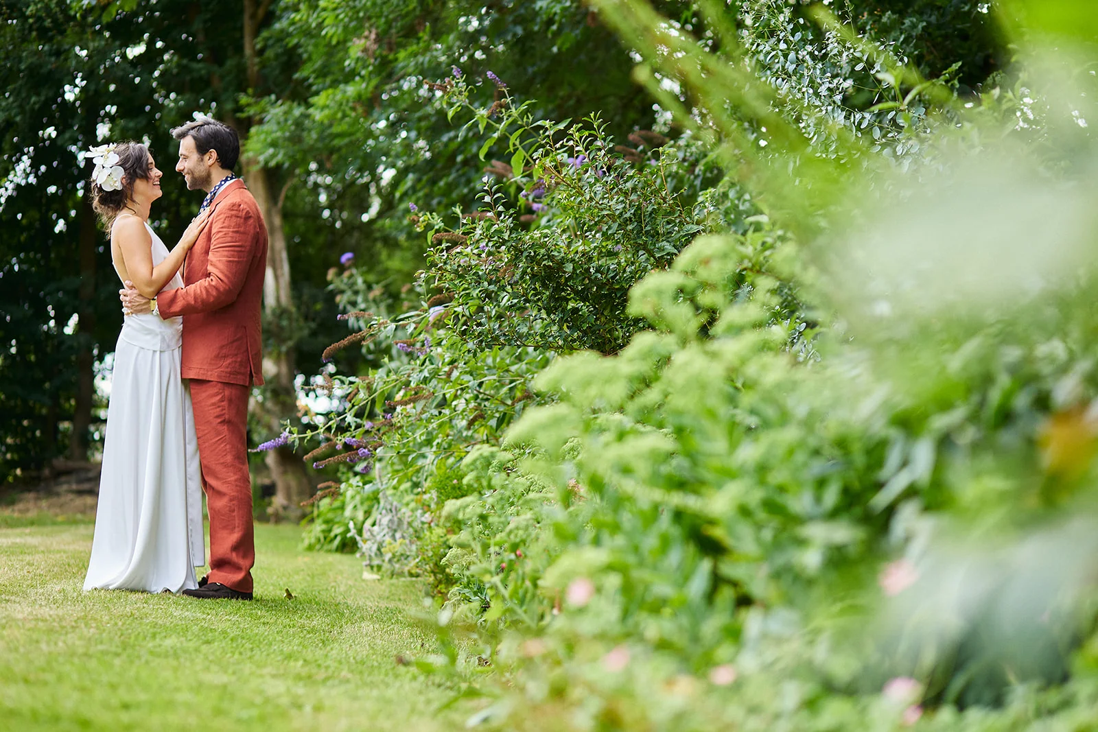 A bride and groom embrace in a lush green garden, framed by foliage in the foreground with the groom wearing a terracotta suit.
