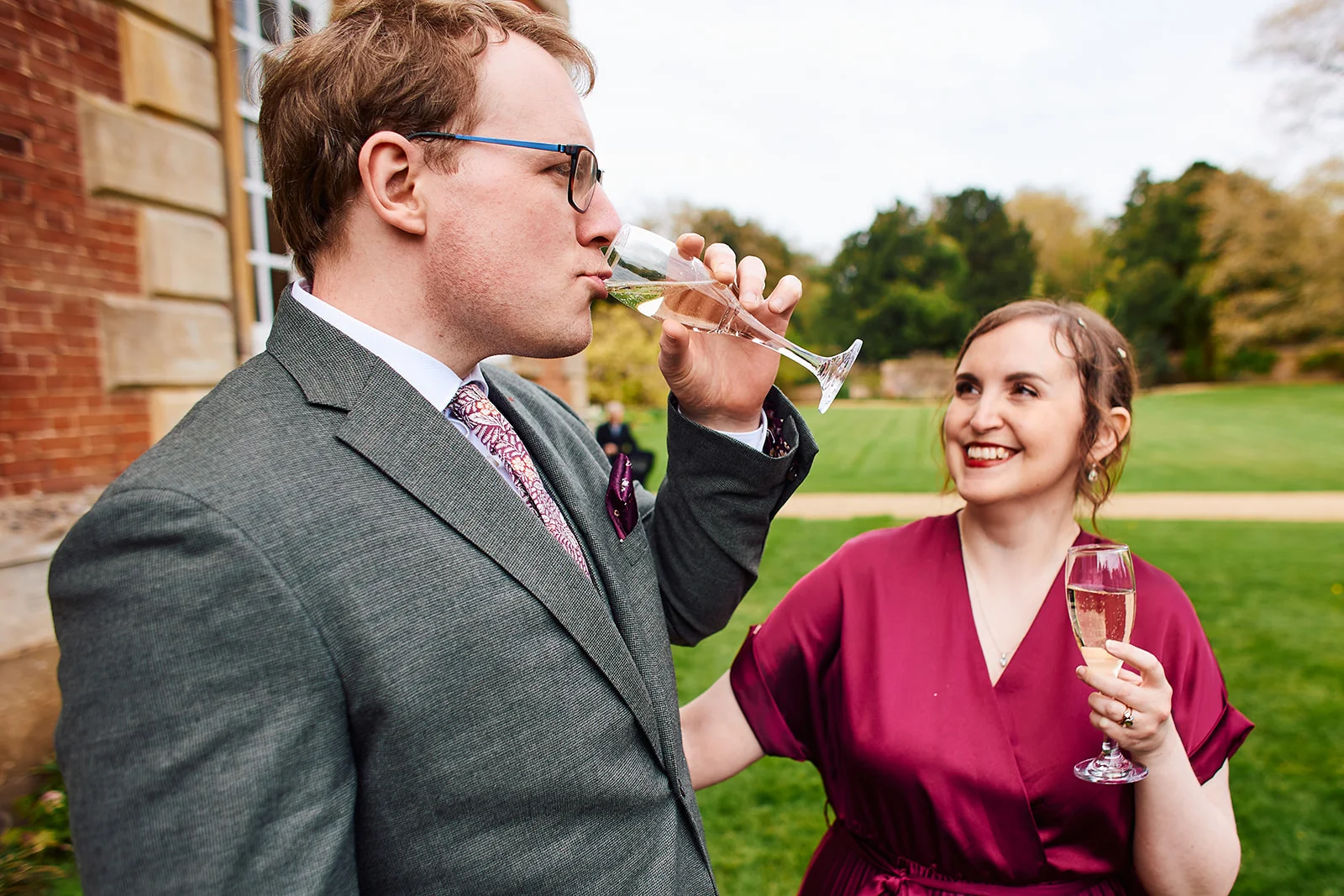 A wedding guest in a grey suit drinks champagne from a flute while smiling at another guest in a burgundy dress holding a glass outdoors.