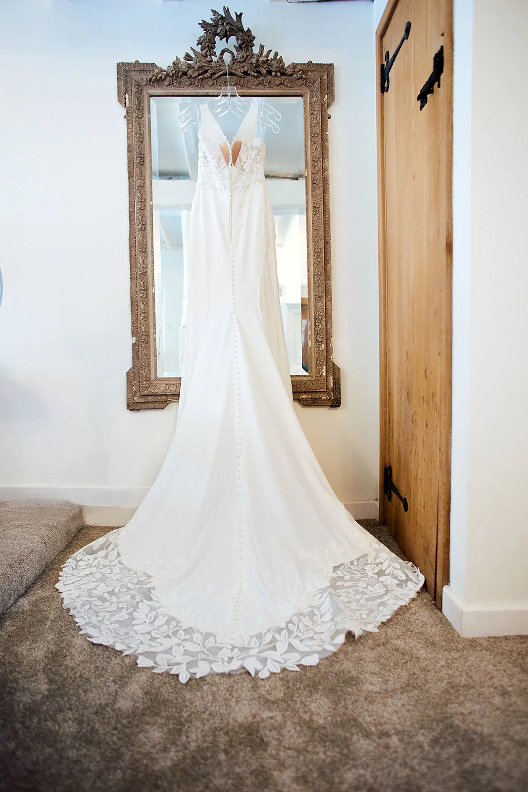 A stunning white lace wedding dress hangs gracefully over a rustic wooden mirror during morning bridal preparations.