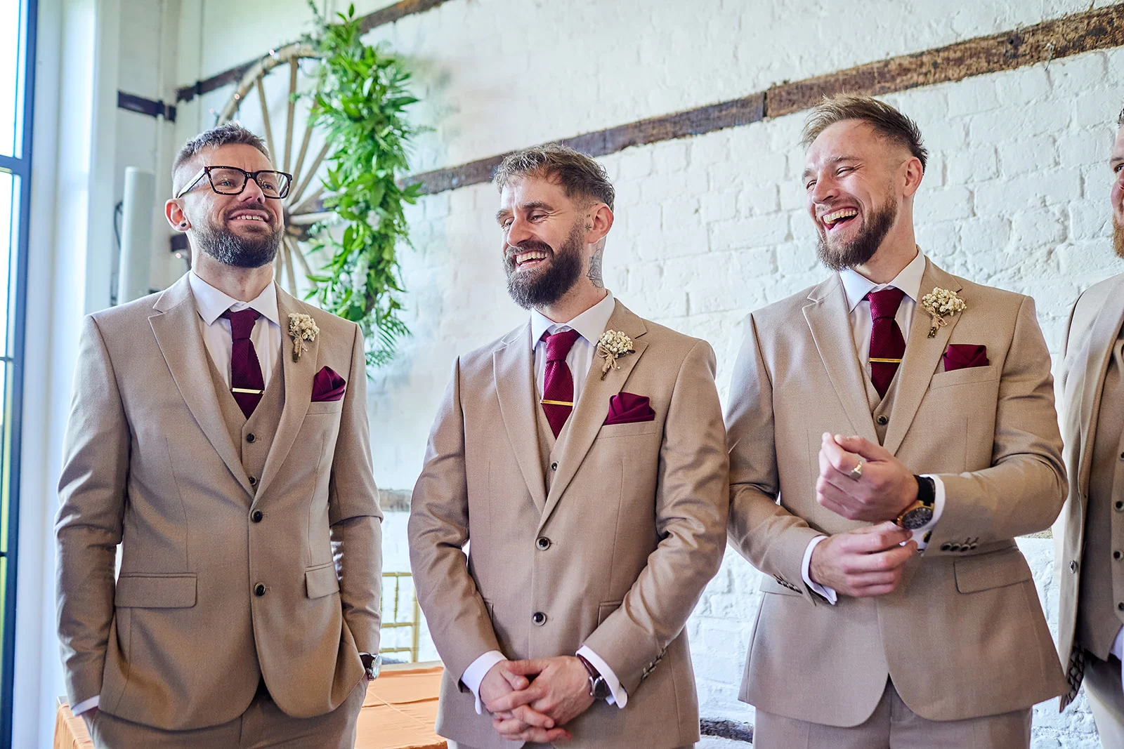 Three smiling groomsmen wearing matching beige three-piece suits and burgundy ties laugh joyfully against a white exposed brick wall backdrop.