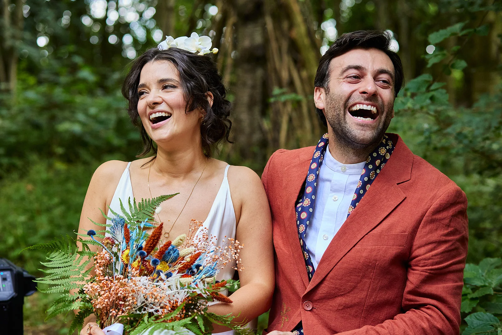 A joyful newlywed couple laugh heartily together outdoors in a lush woodland setting, with the bride holding a colourful textured bouquet containing dried florals and ferns.