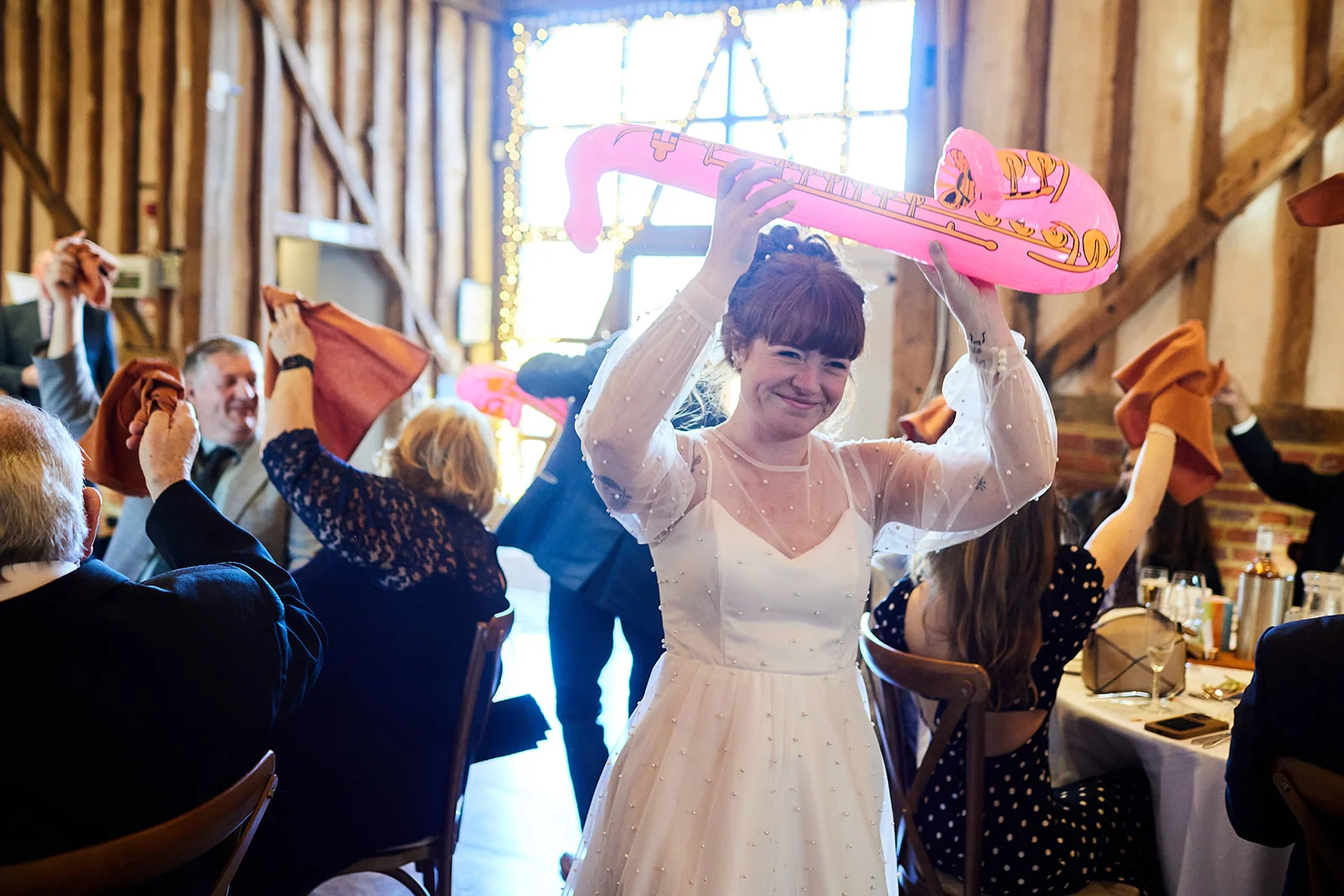 A joyful bride wearing a pearl-embellished gown lifts an inflatable pink saxophone above her head while guests celebrate during wedding speeches inside a rustic barn.