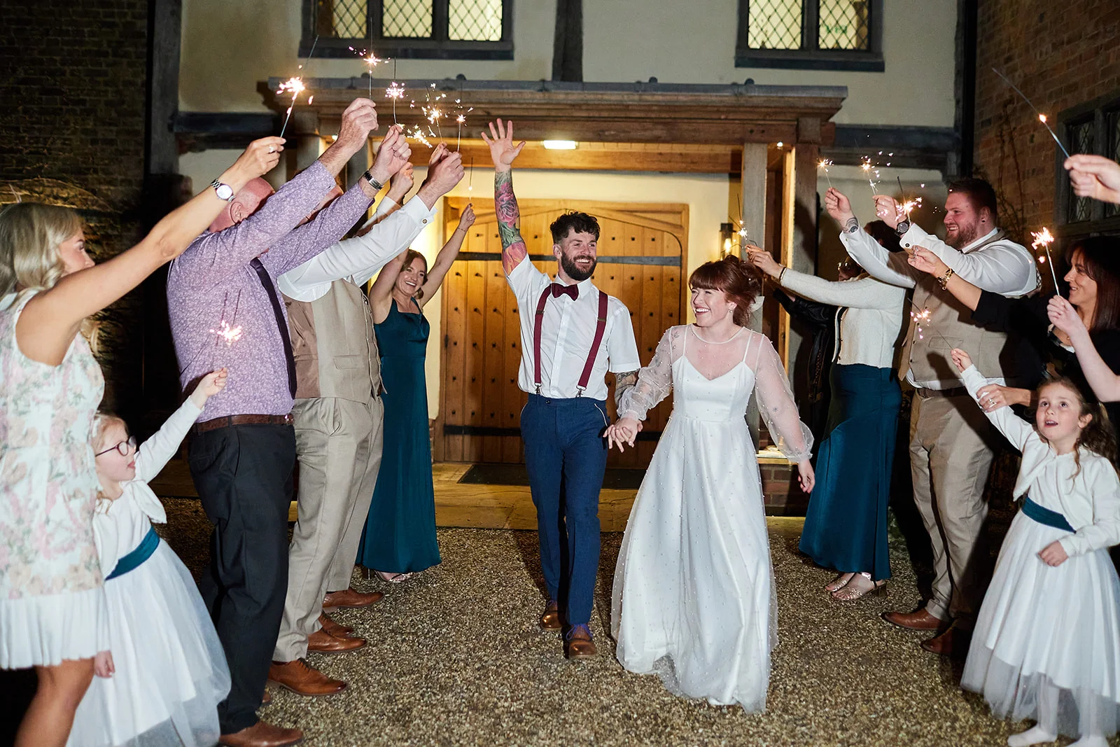 A newlywed couple joyful and laughing walk hand in hand through a sparkling tunnel of lighted sparklers held by cheering guests outside a rustic barn venue at night.