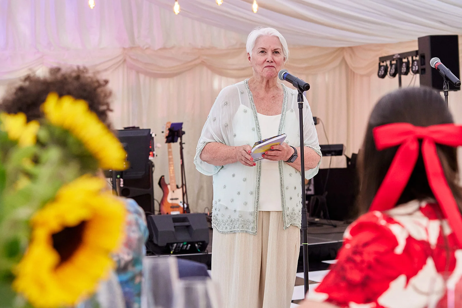 An older woman with short grey hair stands inside a marquee giving a wedding speech into a microphone, holding notes while guests look on.